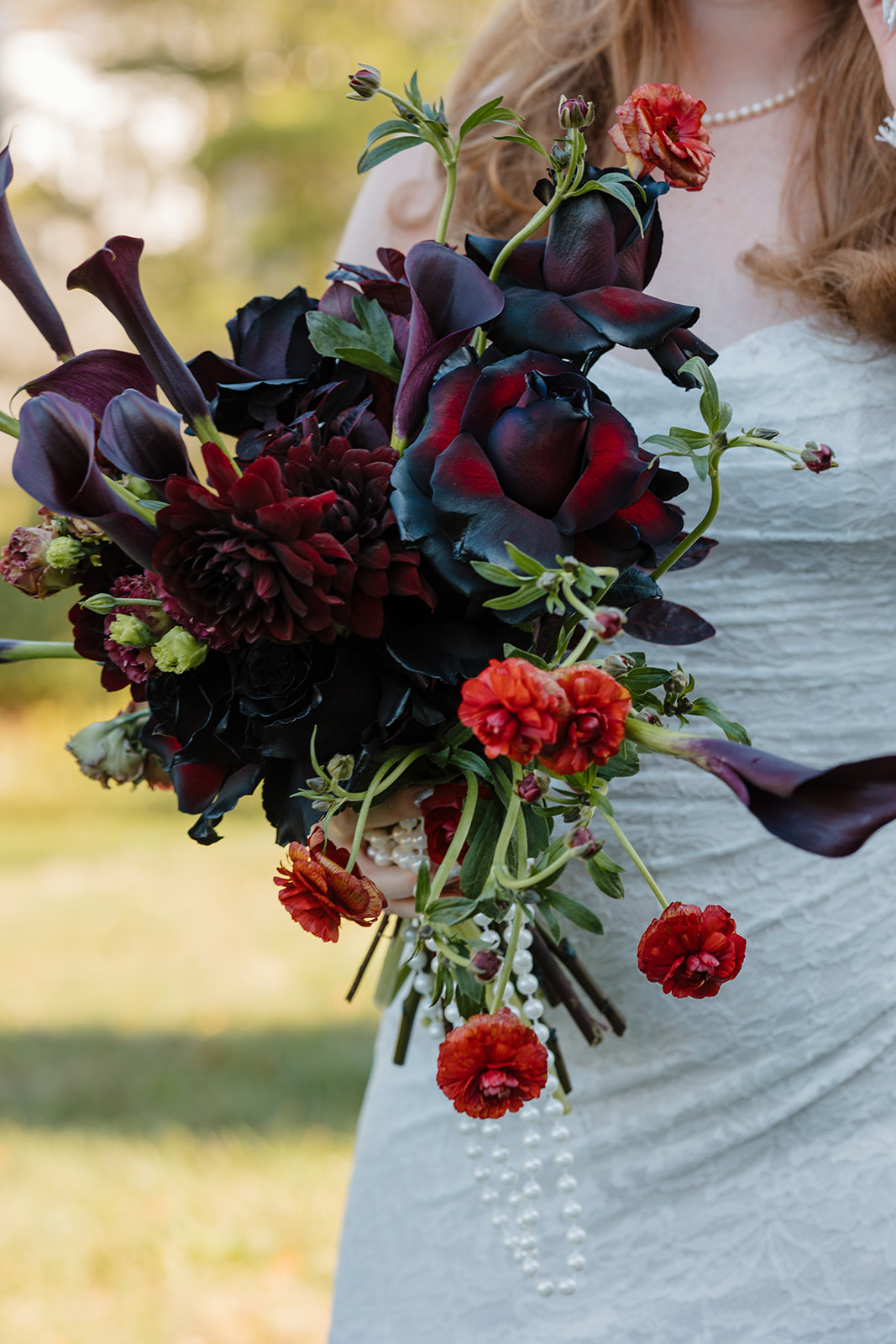 A detail photo of the bride holding her moody wedding bouquet with dark maroon, orange, and red florals and strings of pearls during her fall destination wedding in Maine