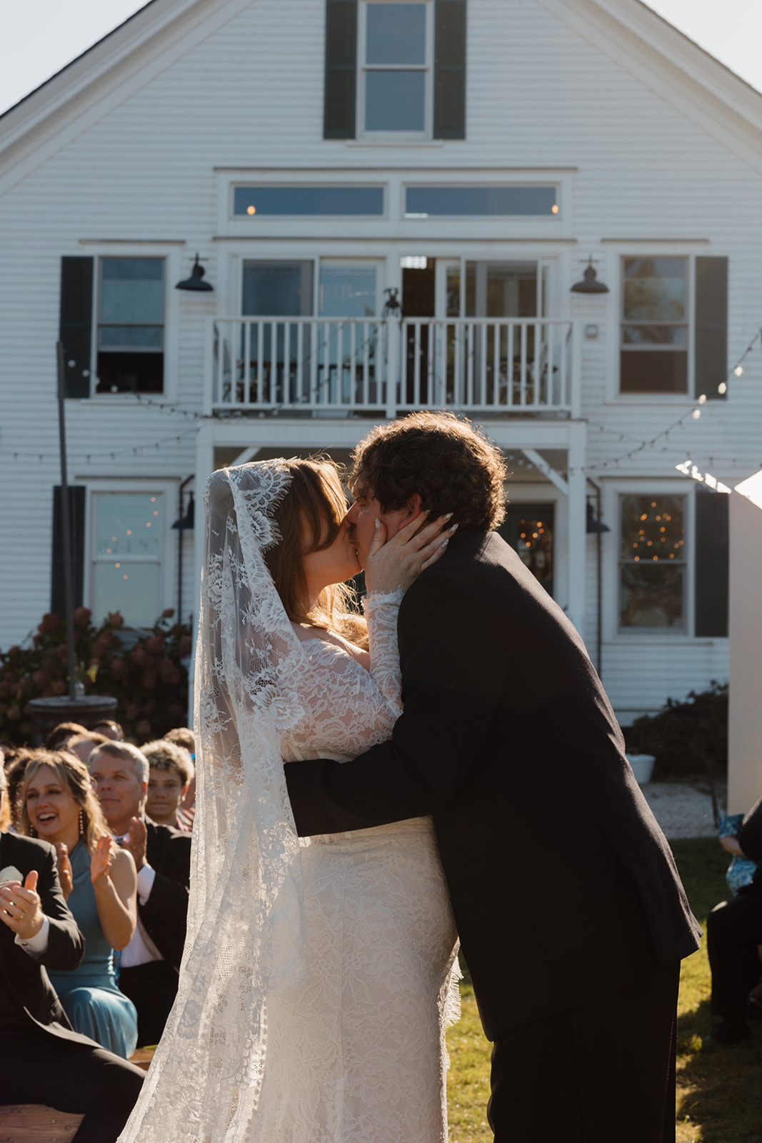 Newlywed couple sharing their first kiss at the altar while guests cheer, set in front of a white coastal New England home