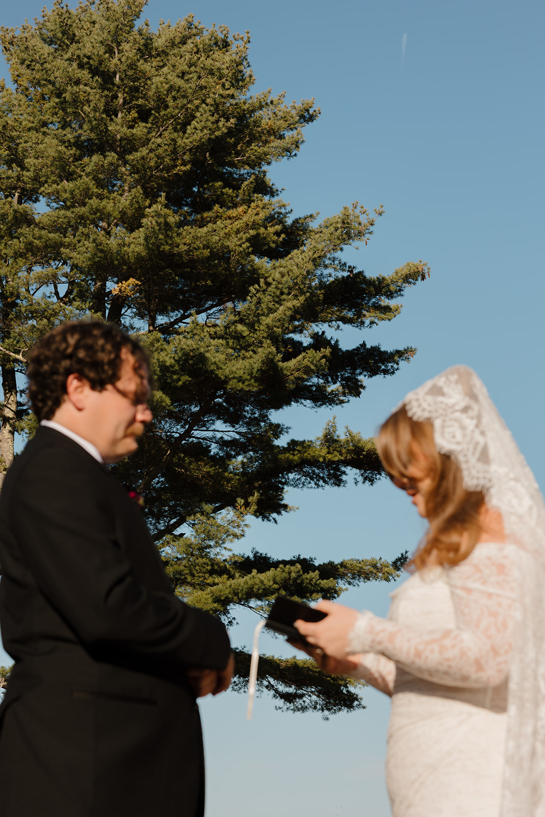Outdoor ceremony moment as the bride reads vows to the groom beneath tall trees and open sky, creating a peaceful and intimate wedding setting