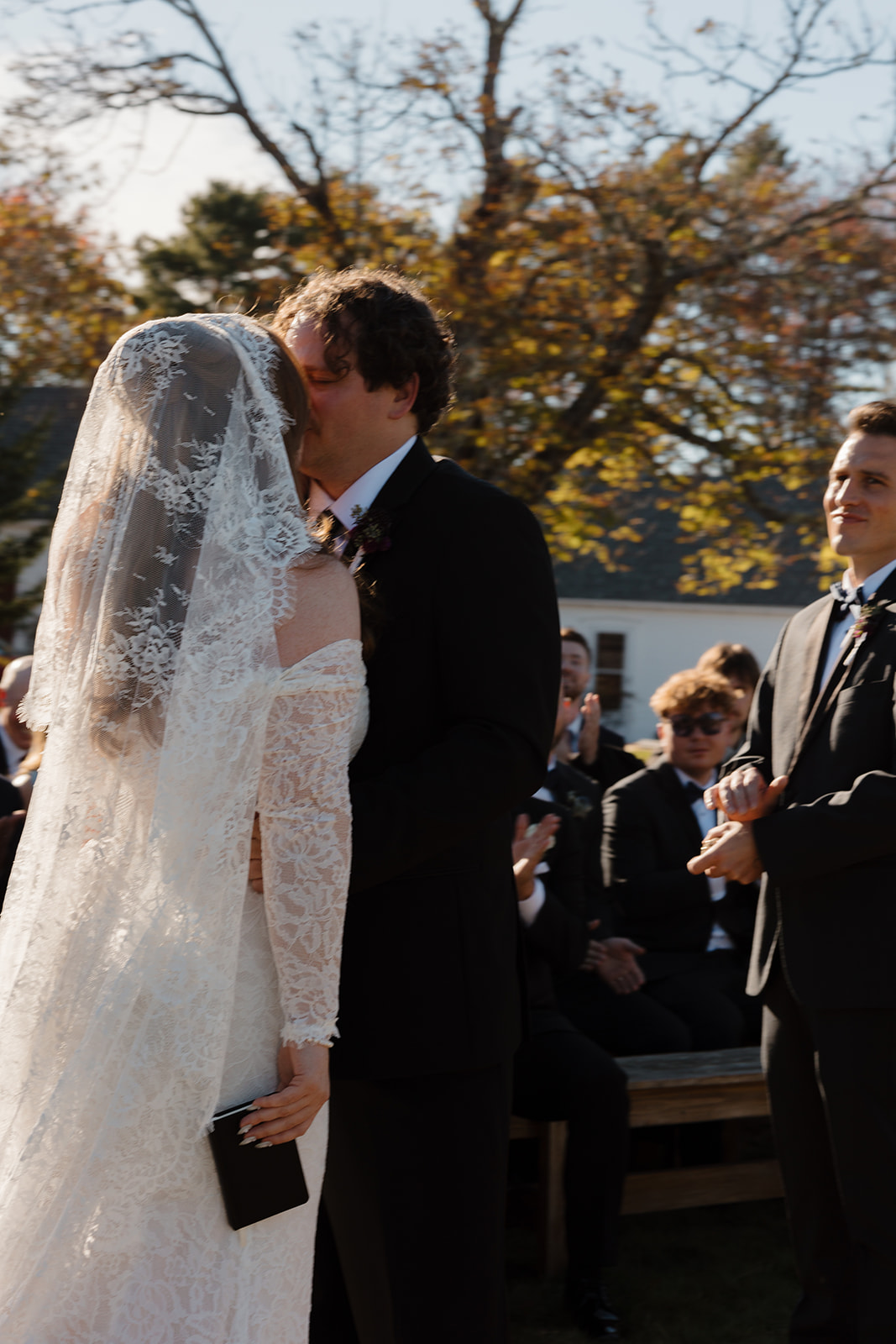 Bride and groom kissing during their outdoor ceremony while guests applaud, surrounded by autumn trees and warm natural light