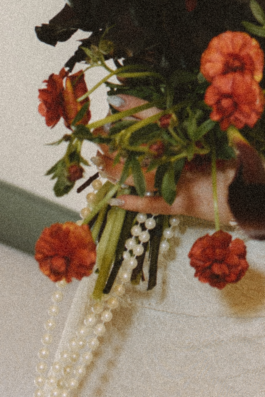 A film detail photo of the bride holding her moody wedding bouquet with dark maroon, orange, and red florals and strings of pearls during her fall destination wedding in Maine