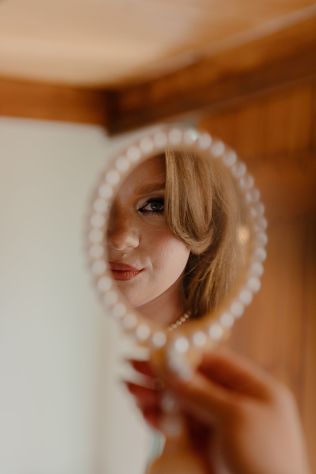 Close-up bridal portrait reflected in a handheld mirror during a Destination Wedding in Maine, highlighting soft makeup, vintage-inspired hair, and pearl details