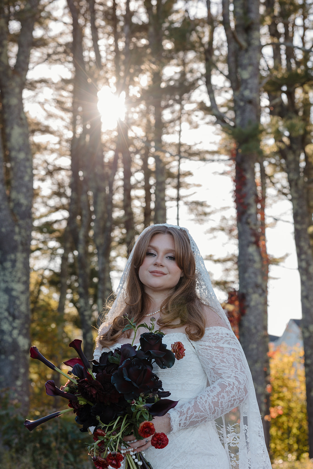 Bride standing beneath tall trees holding her bouquet, sunlight filtering through branches creating a romantic backlit glow