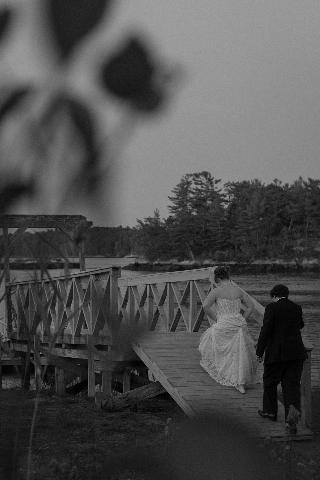 Black and white portrait of the couple crossing a wooden dock together, with coastal marshland and trees in the background
