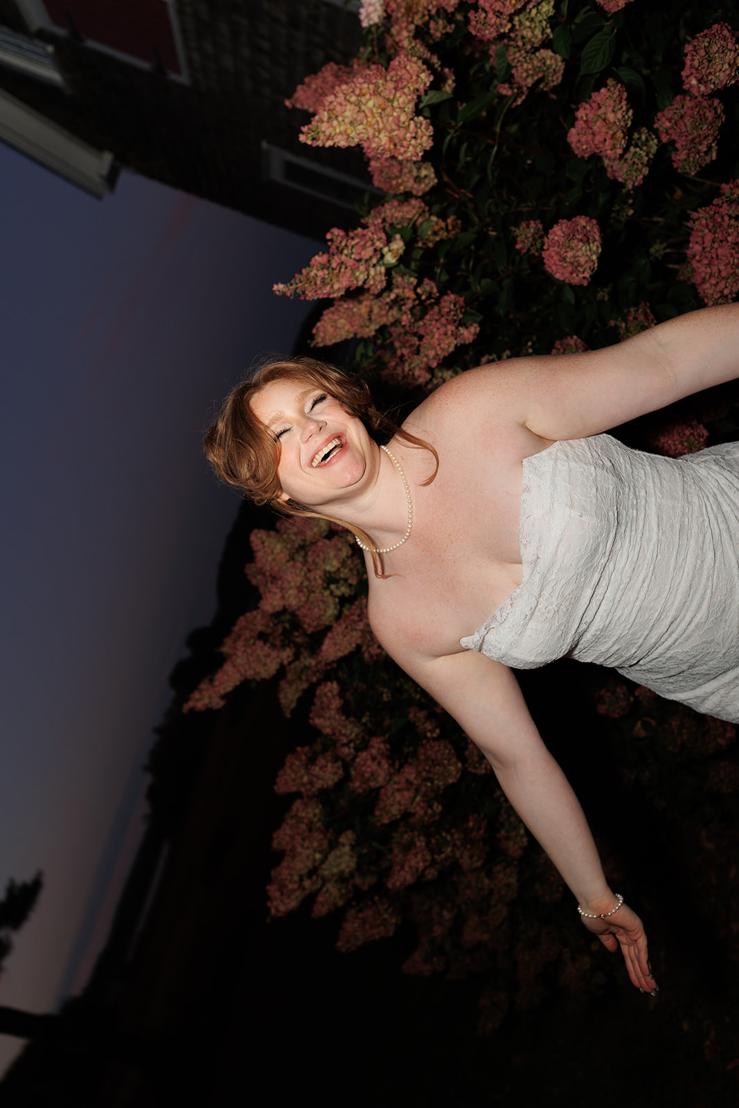 Playful reception portrait of the bride laughing while leaning back near pink hydrangea bushes at dusk
