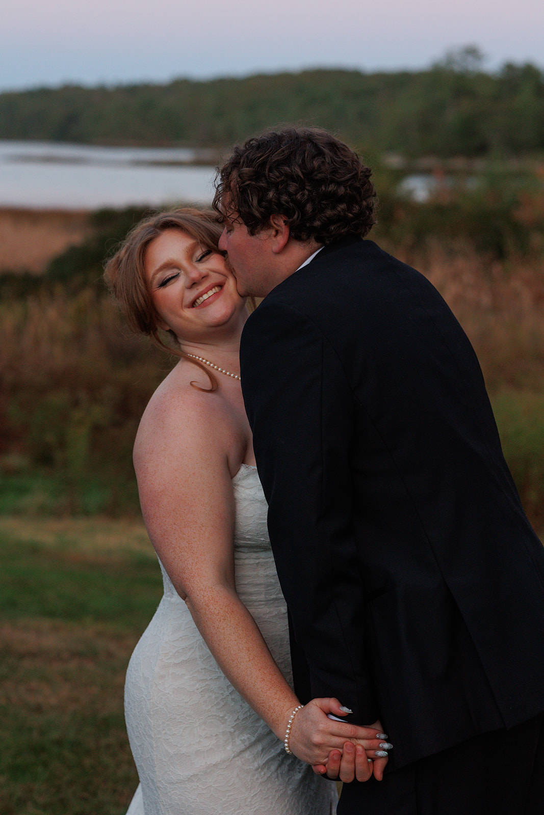 Romantic portrait of the groom kissing the bride’s cheek outdoors, with soft greenery and water in the background during golden hour