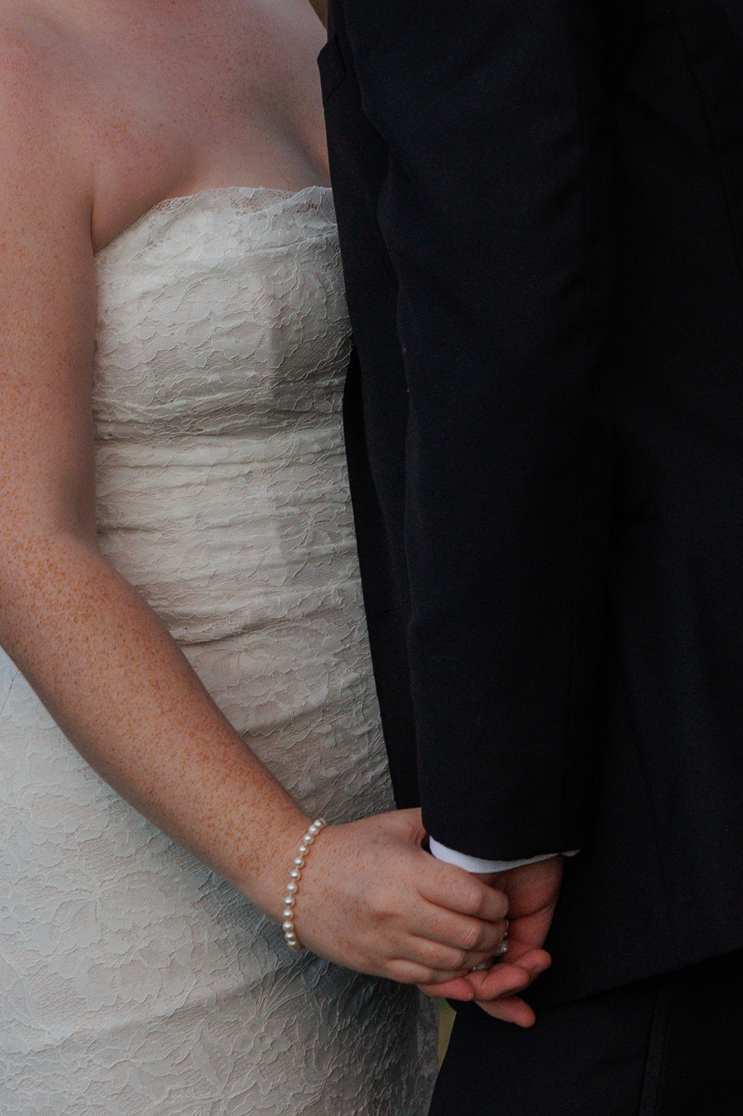 Close-up of the bride holding the groom’s hand from behind during a Destination Wedding in Maine, showcasing her lace strapless gown and pearl bracelet