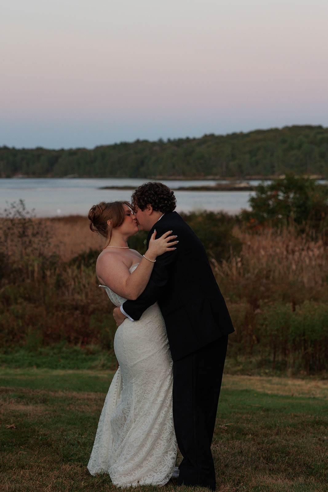 Romantic waterfront kiss at blue hour as the bride and groom hold each other close, surrounded by tall grass and soft pastel sky