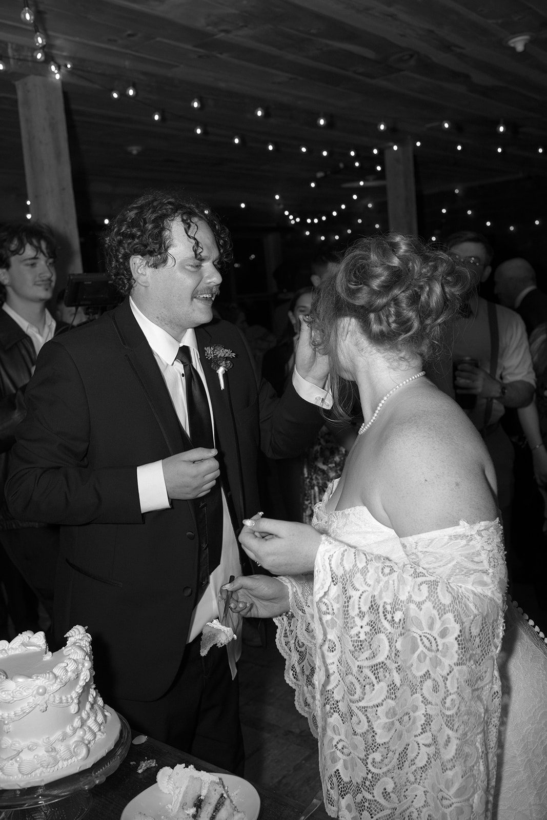 Black and white reception moment as the groom gently wipes frosting from the bride’s face while guests watch and laugh
