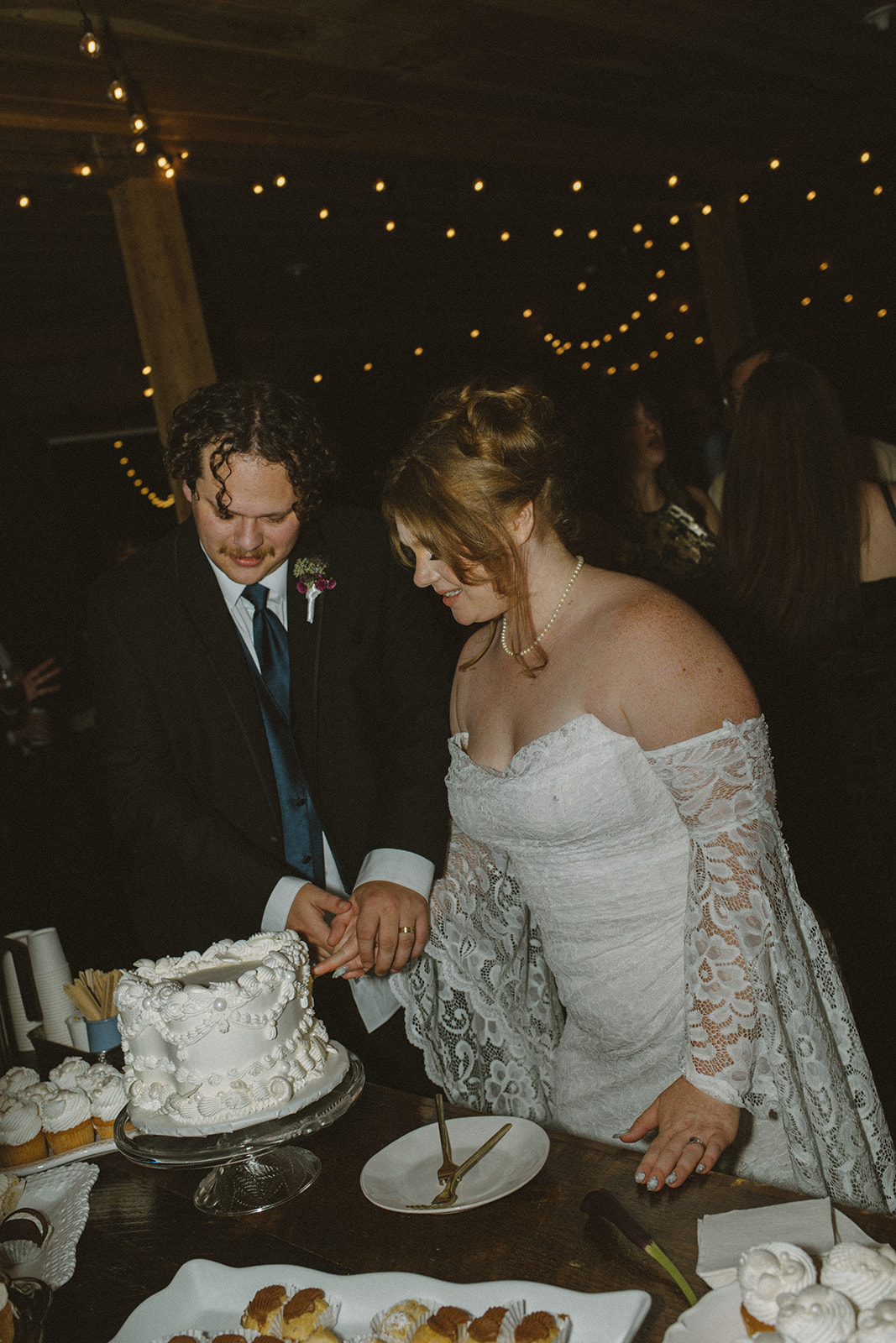 Bride and groom cutting their wedding cake together during a Destination Wedding in Maine, surrounded by candlelight, desserts, and warm string lights inside a rustic reception space