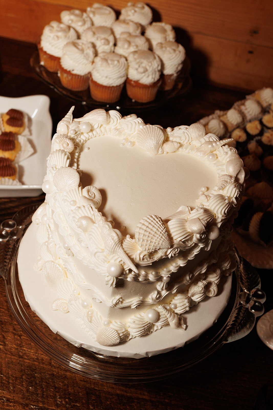 A elegant white wedding cake in the shape of a heart with seashells and pearls decorating it, at an elegant and moody destination wedding in Maine.
