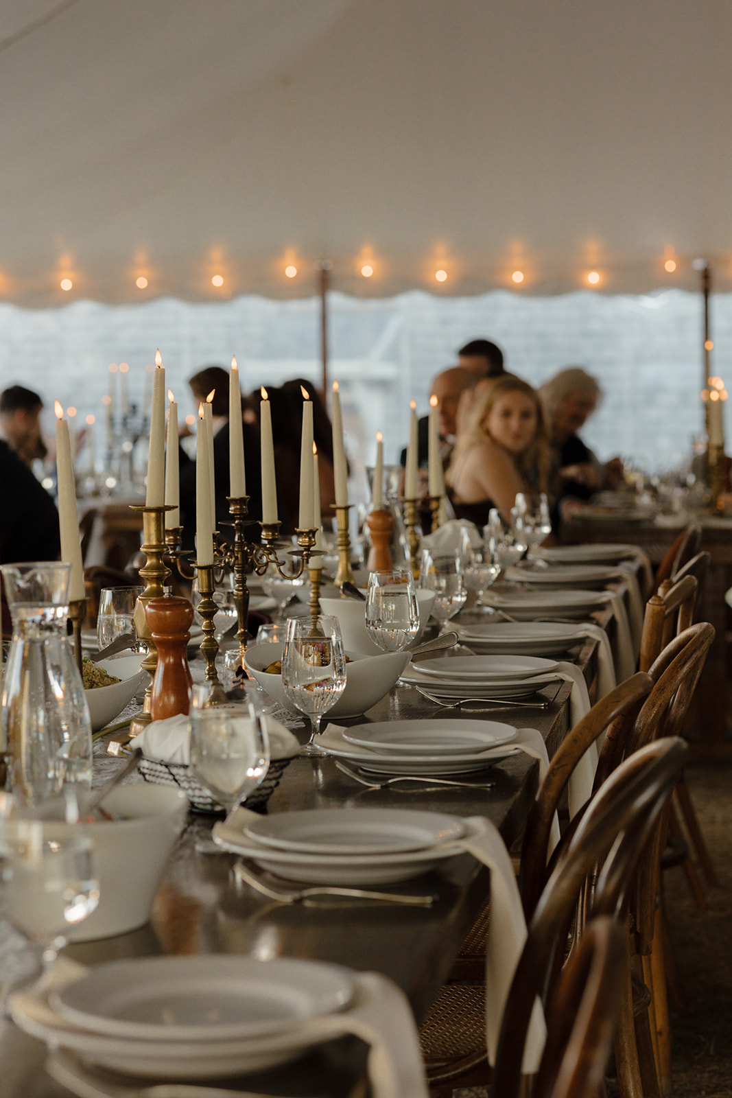 An elegant tablescape with bronze candle holder and candles, brown wood chairs, white plates and napkins, and lights around the tables handing on the tent in the background.