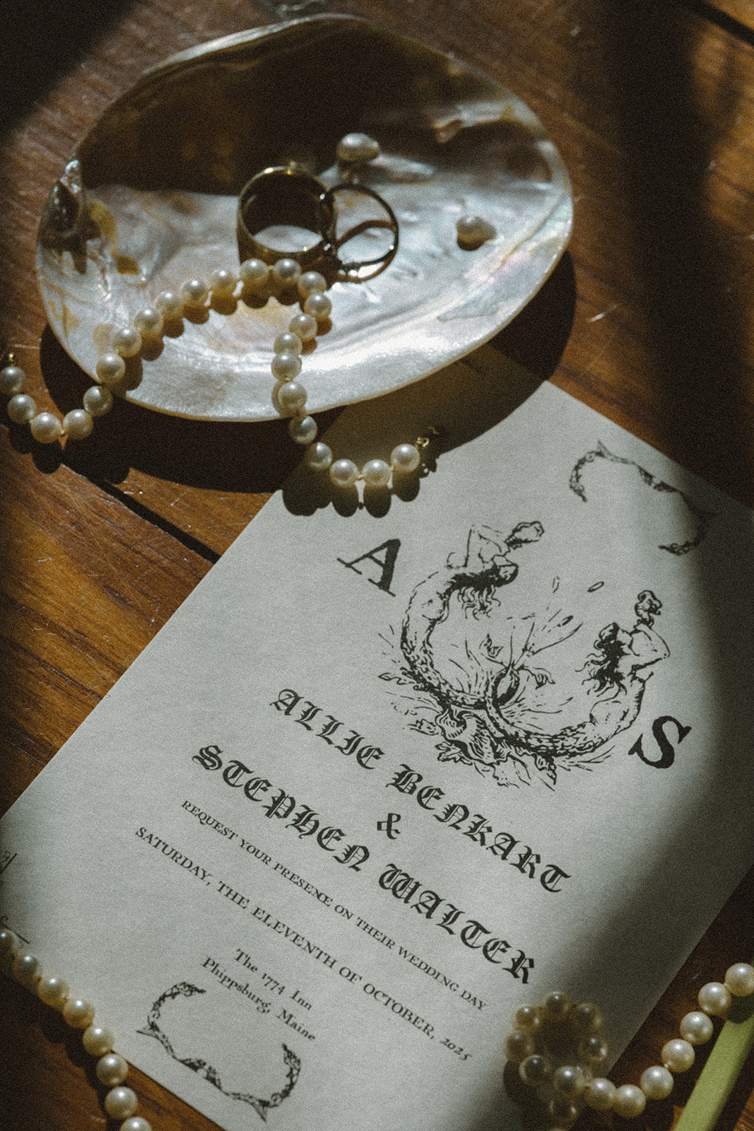 An elegant flatlay detail of the couples destination wedding in Maine wedding invite, strings of pearls, and their wedding rings on a pearl ring holder tray.