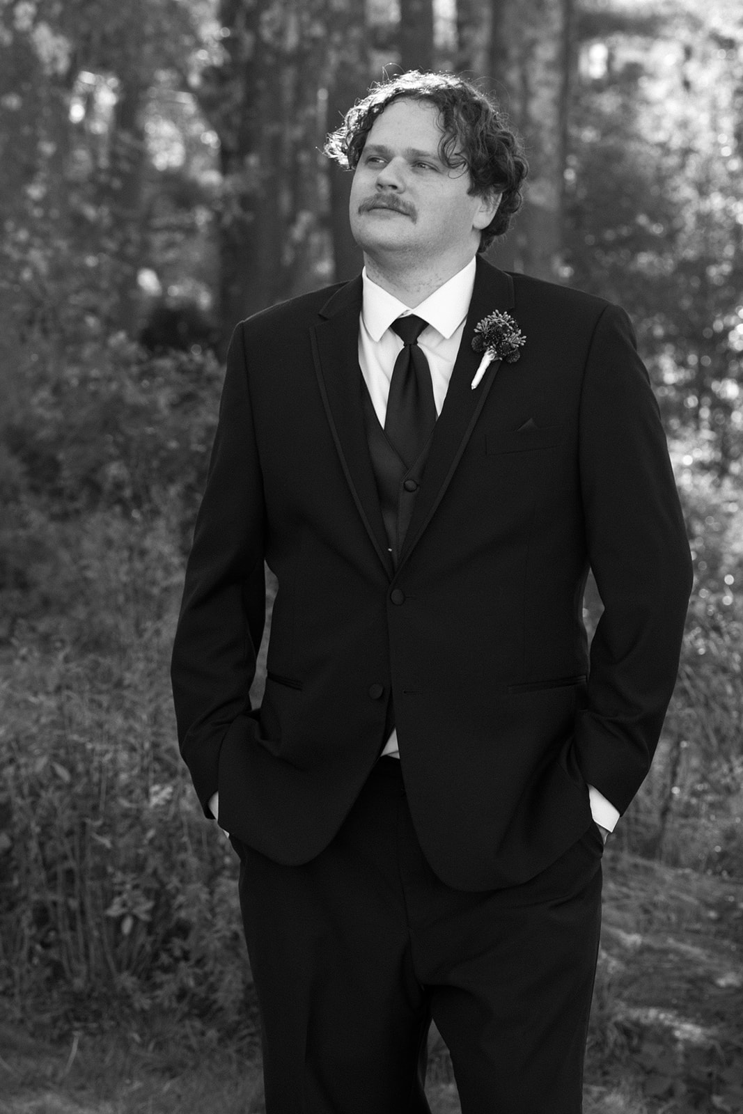 Groom portrait standing outdoors in a black suit and tie during a Destination Wedding in Maine, hands in pockets surrounded by tall coastal trees
