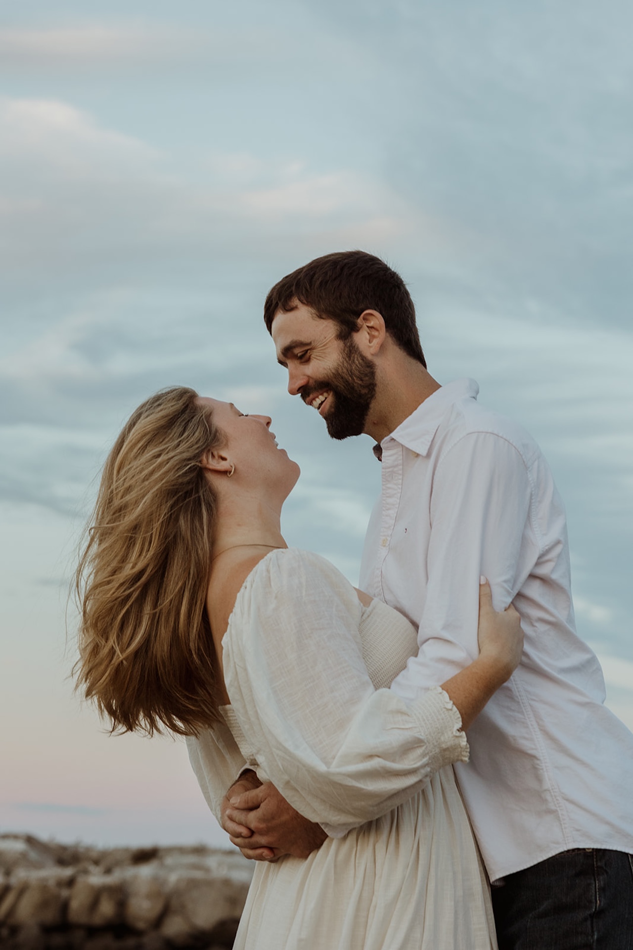 Romantic candid engagement portrait of couple laughing together while embracing on rocky beach coastline.