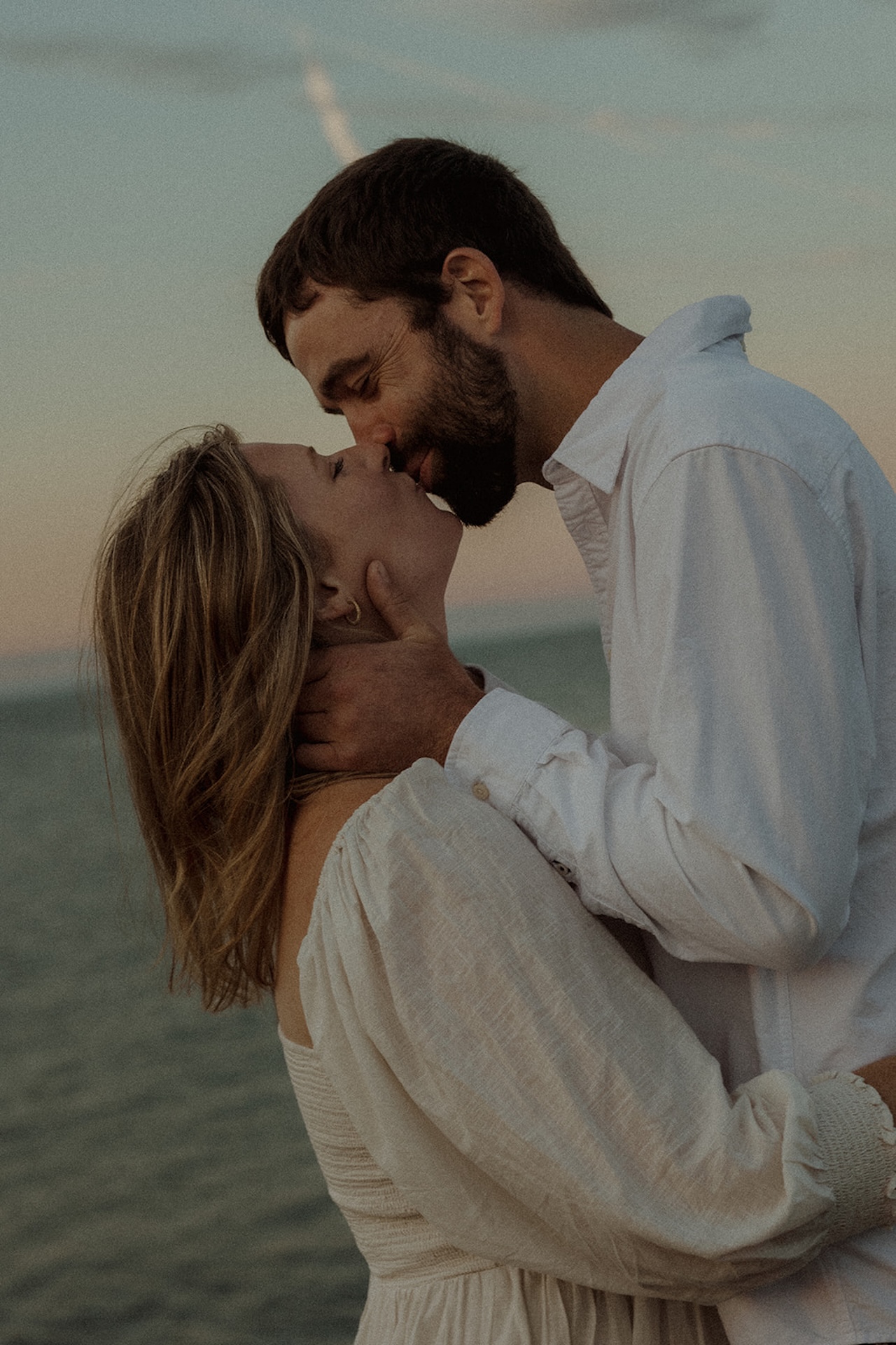 Close-up engagement photo of couple kissing along the shoreline at Nubble Lighthouse in York Maine during soft pastel sunset light.