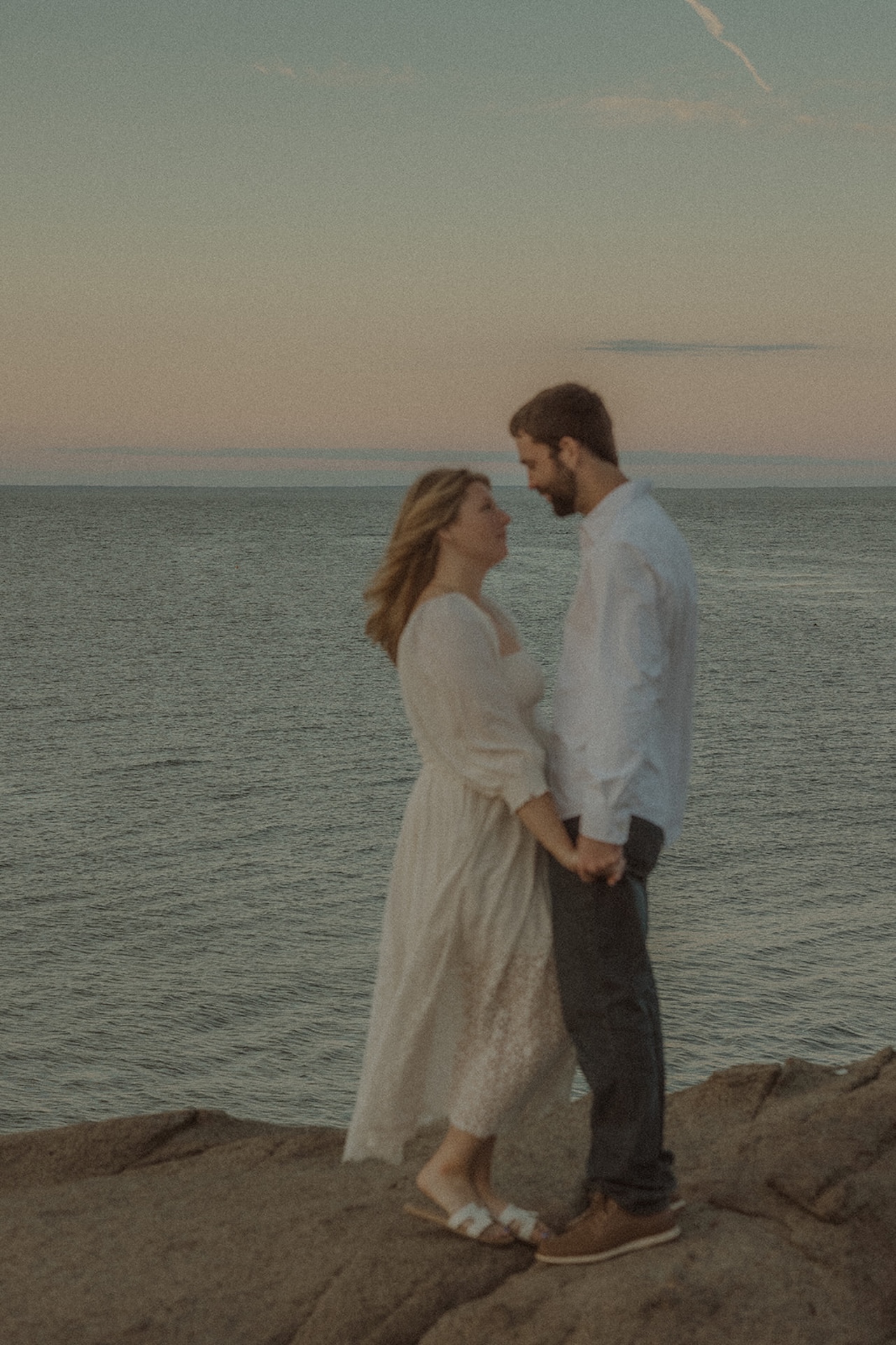 Soft, candid engagement image of couple holding hands while standing on coastal rocks overlooking the ocean.