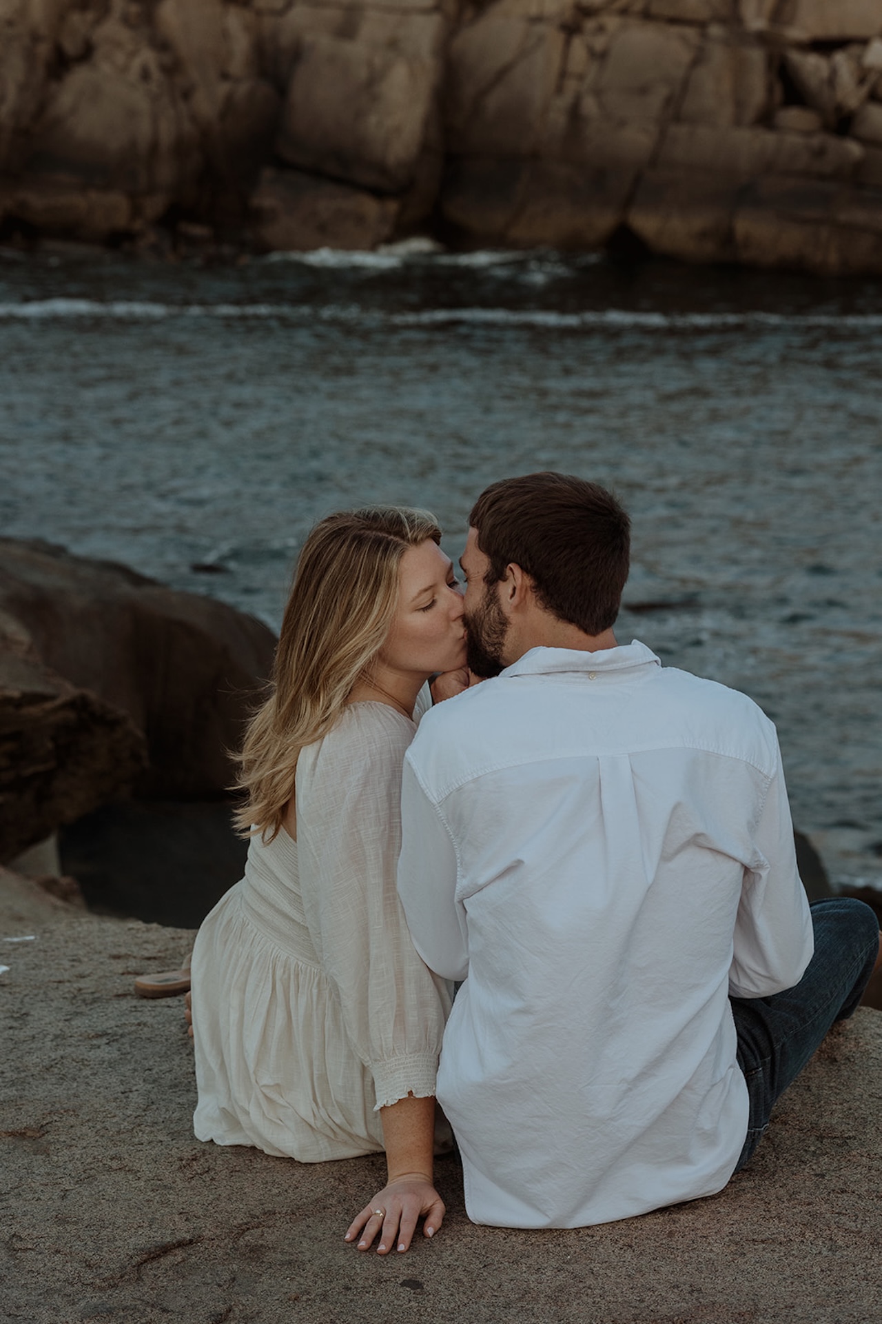 Romantic seaside engagement photo of couple sitting on rocks kissing near Nubble Lighthouse in York Maine with waves behind them.