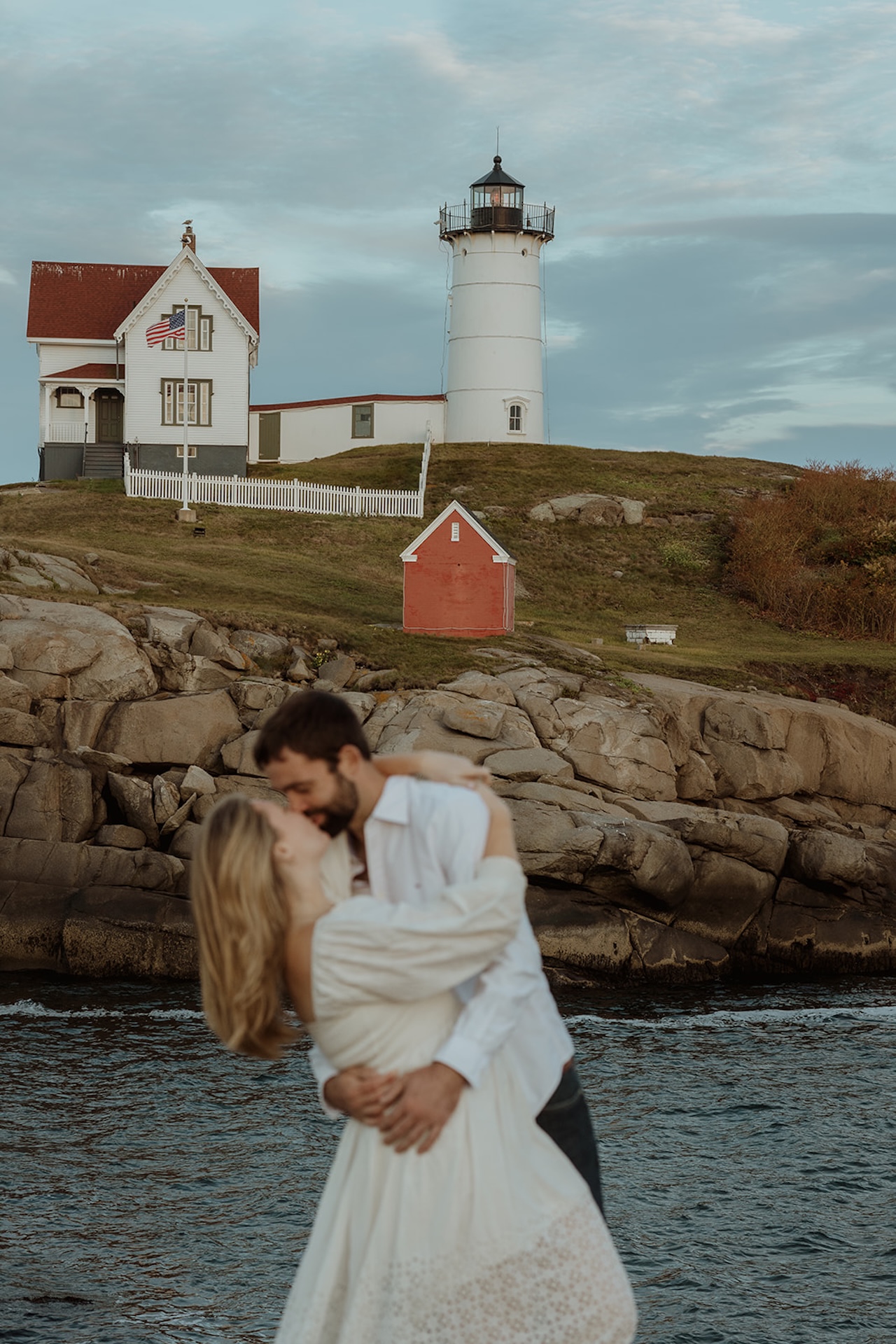 Wide scenic image of Nubble Lighthouse in York Maine overlooking the rocky coast while the couple embraces in the foreground during their engagement session.