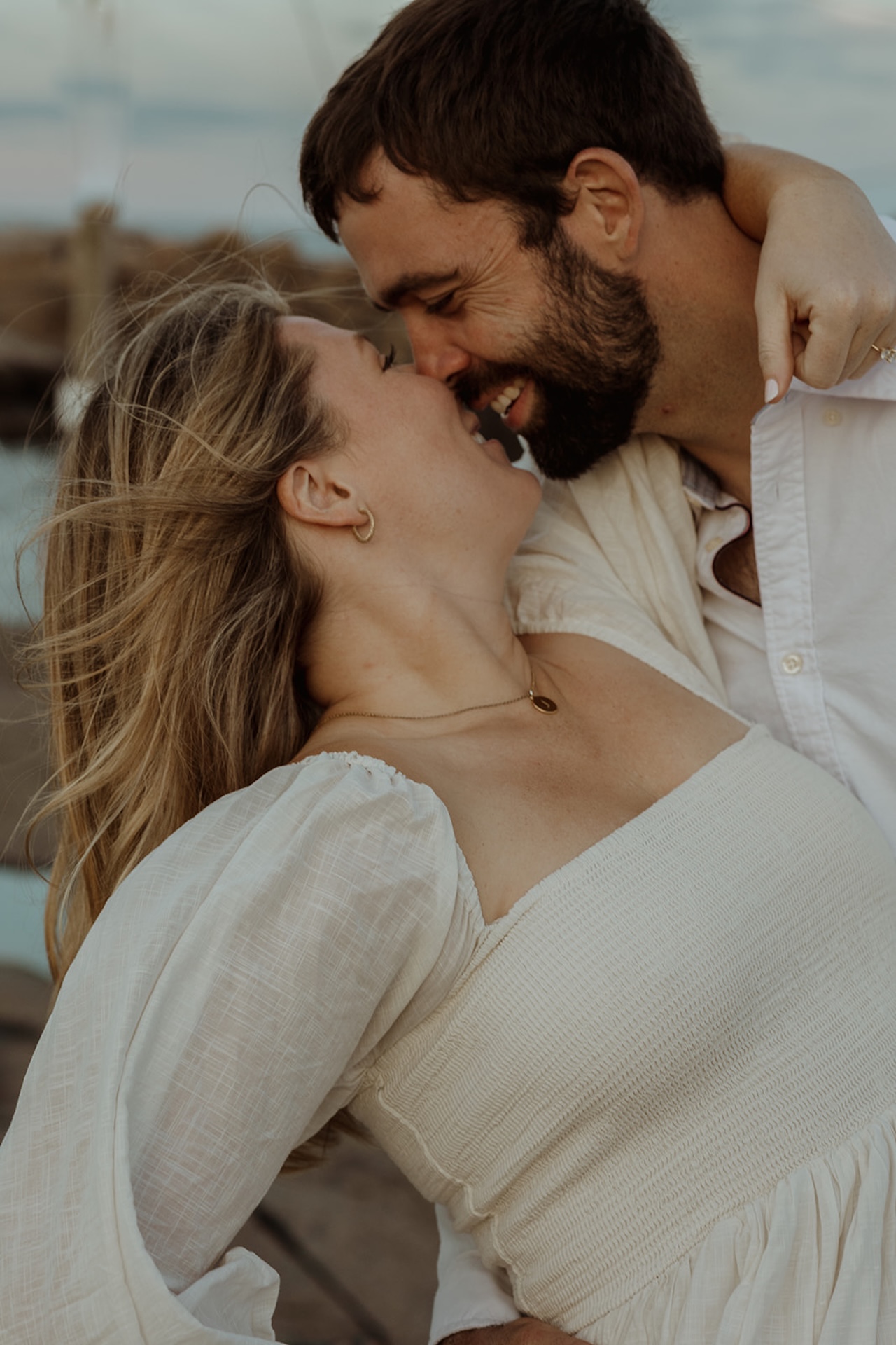 Romantic engagement photo of couple kissing in front of a coastal lighthouse on a rocky shoreline at sunset.