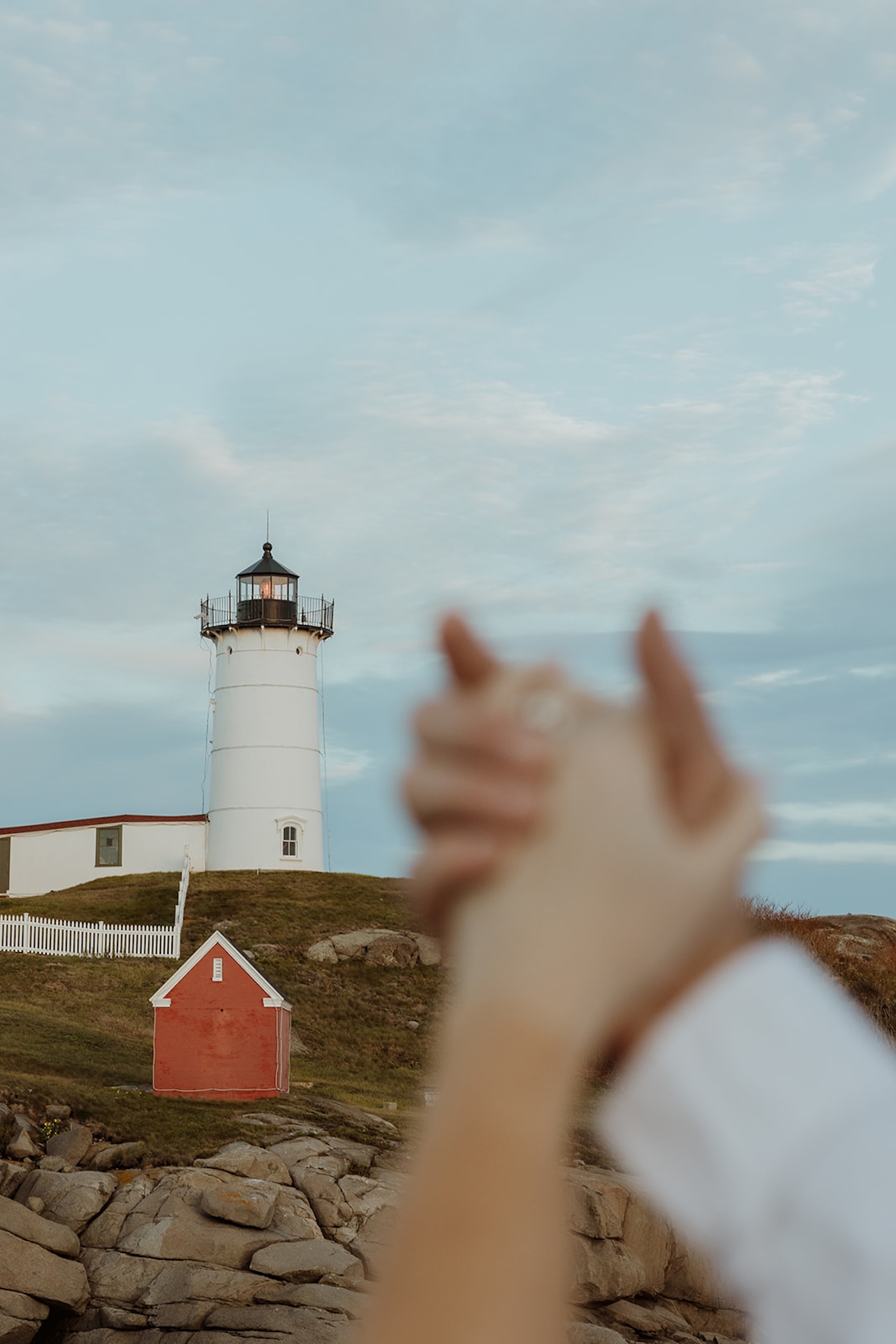 Romantic engagement photo of couple holding hands in front of a coastal lighthouse on a rocky shoreline at sunset.