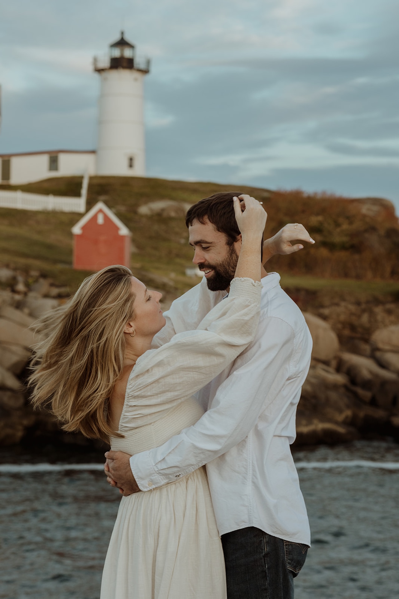Wide scenic image of Nubble Lighthouse in York Maine overlooking the rocky coast while the couple embraces in the foreground during their engagement session.