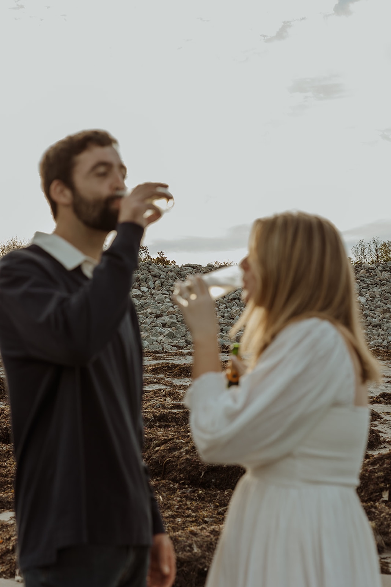 Blurry, golden hour engagement photo of couple sipping champagne together near Nubble Lighthouse in York Maine with rocky coastline behind them.