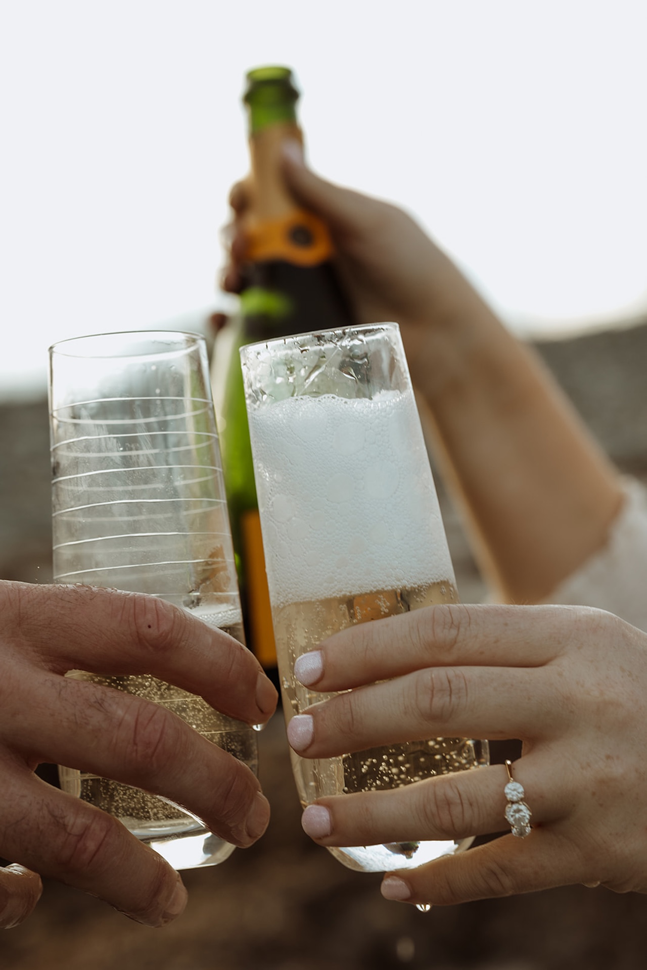 Candid photo of a couple clinking champagne glasses with the sunset behind them during their engagemen shoot at Nubble Lighthouse in York Maine
