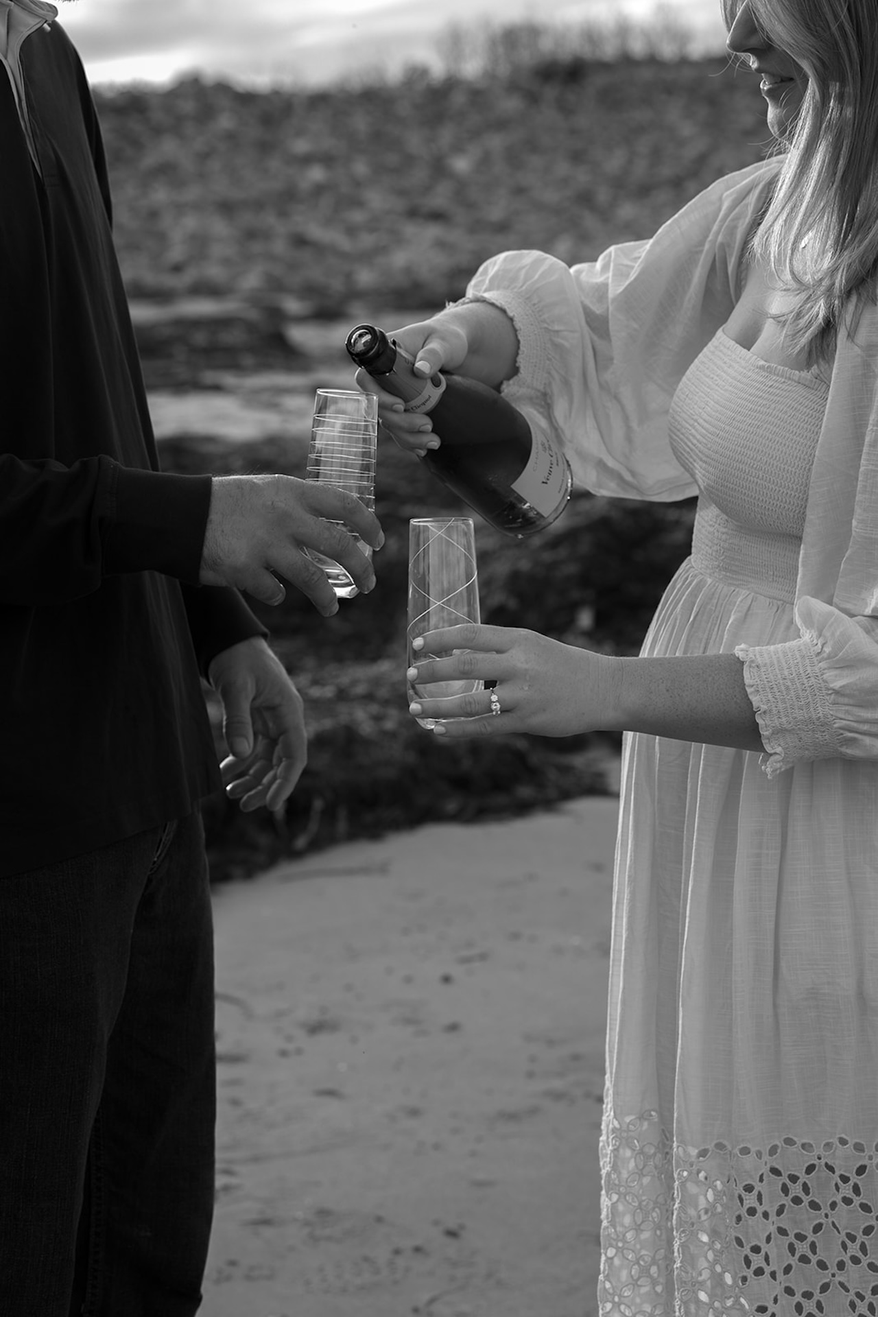Bride-to-be pouring champagne into two glasses during a romantic coastal engagement session, her engagement ring visible as they celebrate by the ocean.