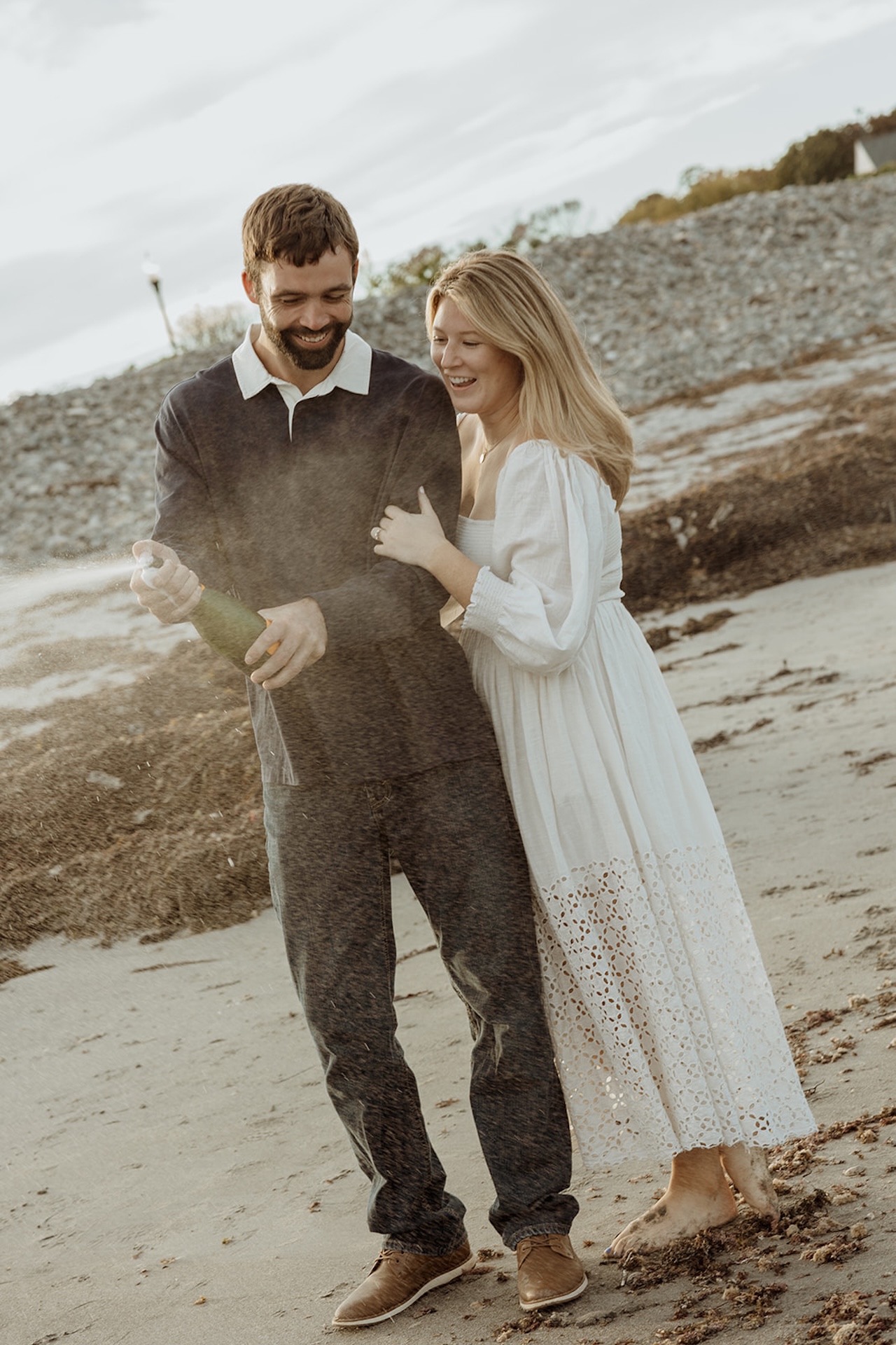 Couple popping champagne on the beach during their engagement session at Nubble Lighthouse in York Maine with seaweed-covered sand and rocky shoreline in the background.