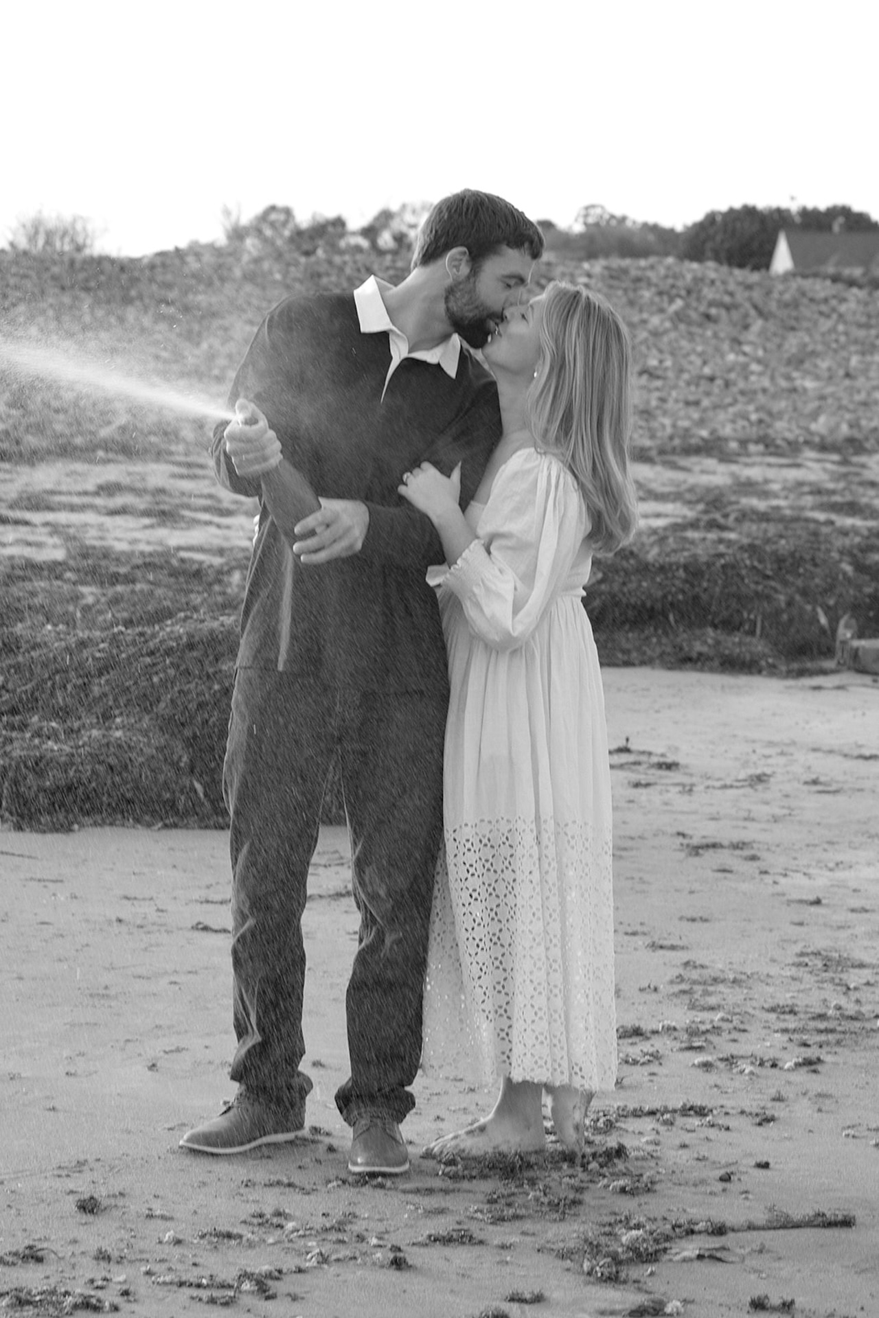 Black and white photo of a couple popping champagne on the beach during their engagement session at Nubble Lighthouse in York Maine with seaweed-covered sand and rocky shoreline in the background.