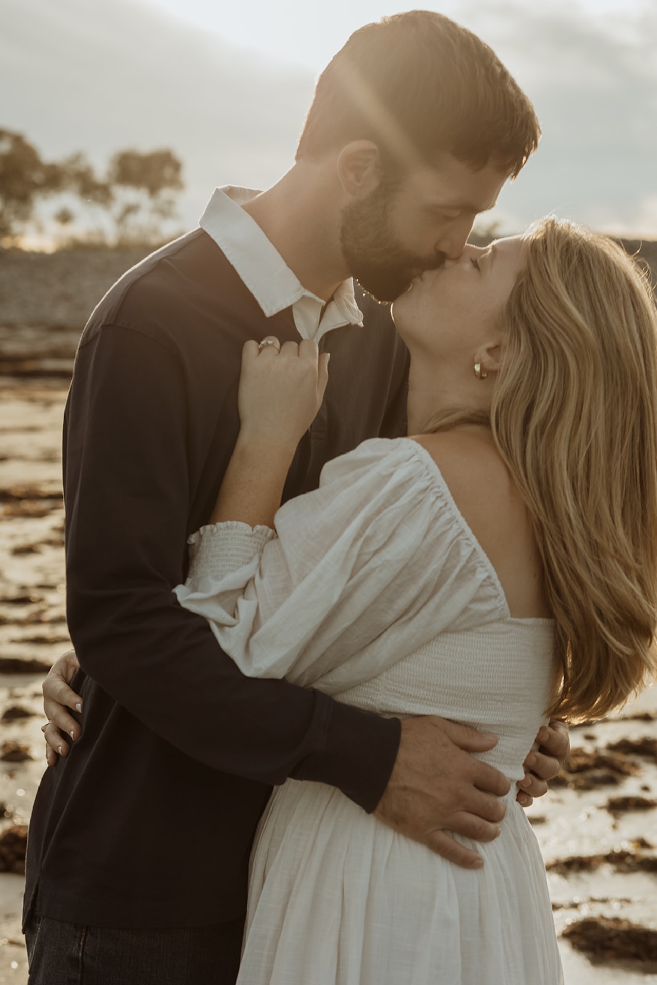 Couple popping champagne on the beach during their engagement session at Nubble Lighthouse in York Maine with seaweed-covered sand and rocky shoreline in the background.