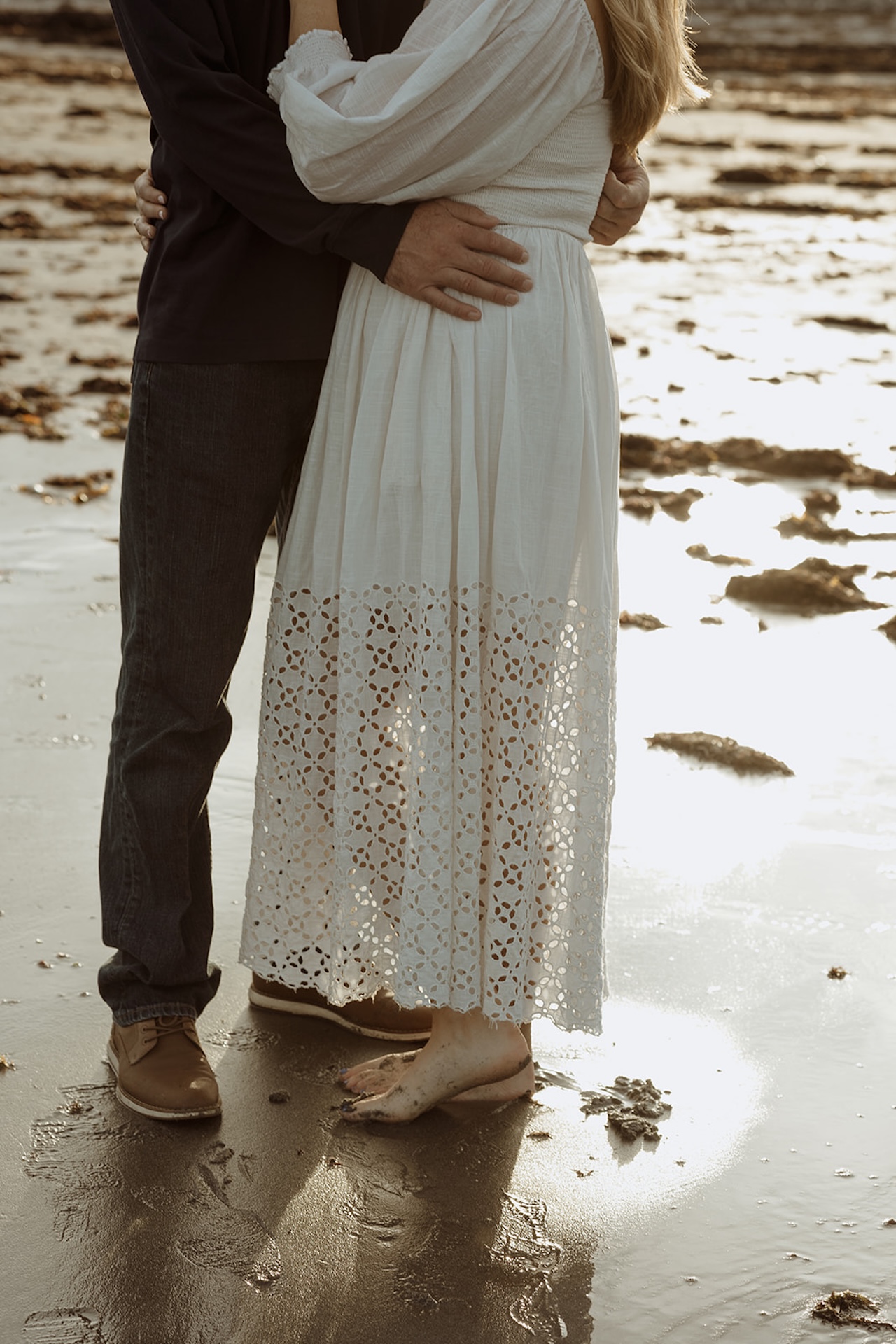 Close detail of couple embracing on the beach with textured white dress and wet sand reflecting golden light.