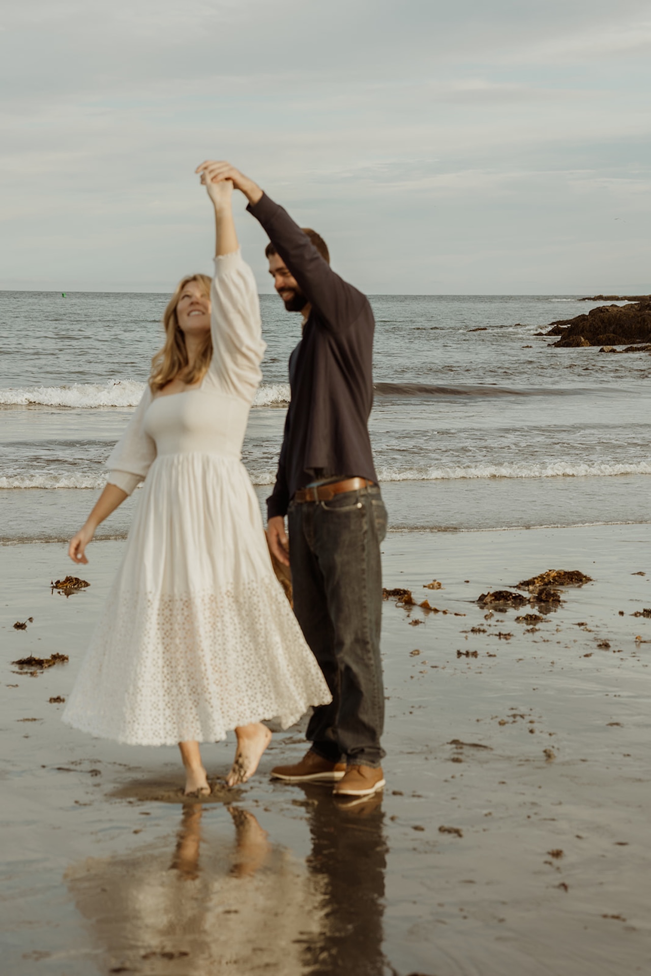Playful engagement photo of couple twirling barefoot in the sand along the shoreline at sunset.