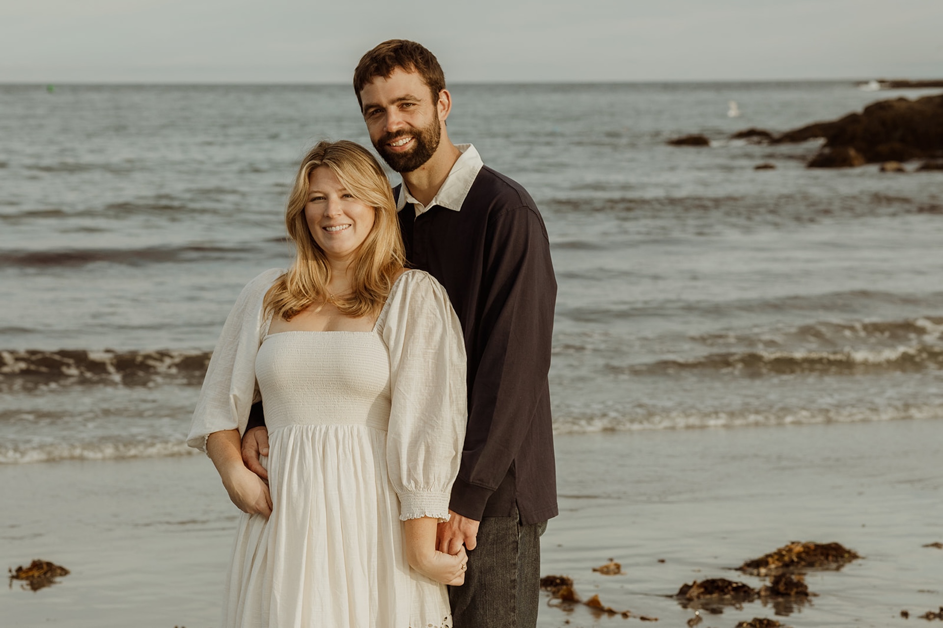 Beach engagement photo of couple smiling at camera with ocean waves behind them near Nubble Lighthouse in York Maine.