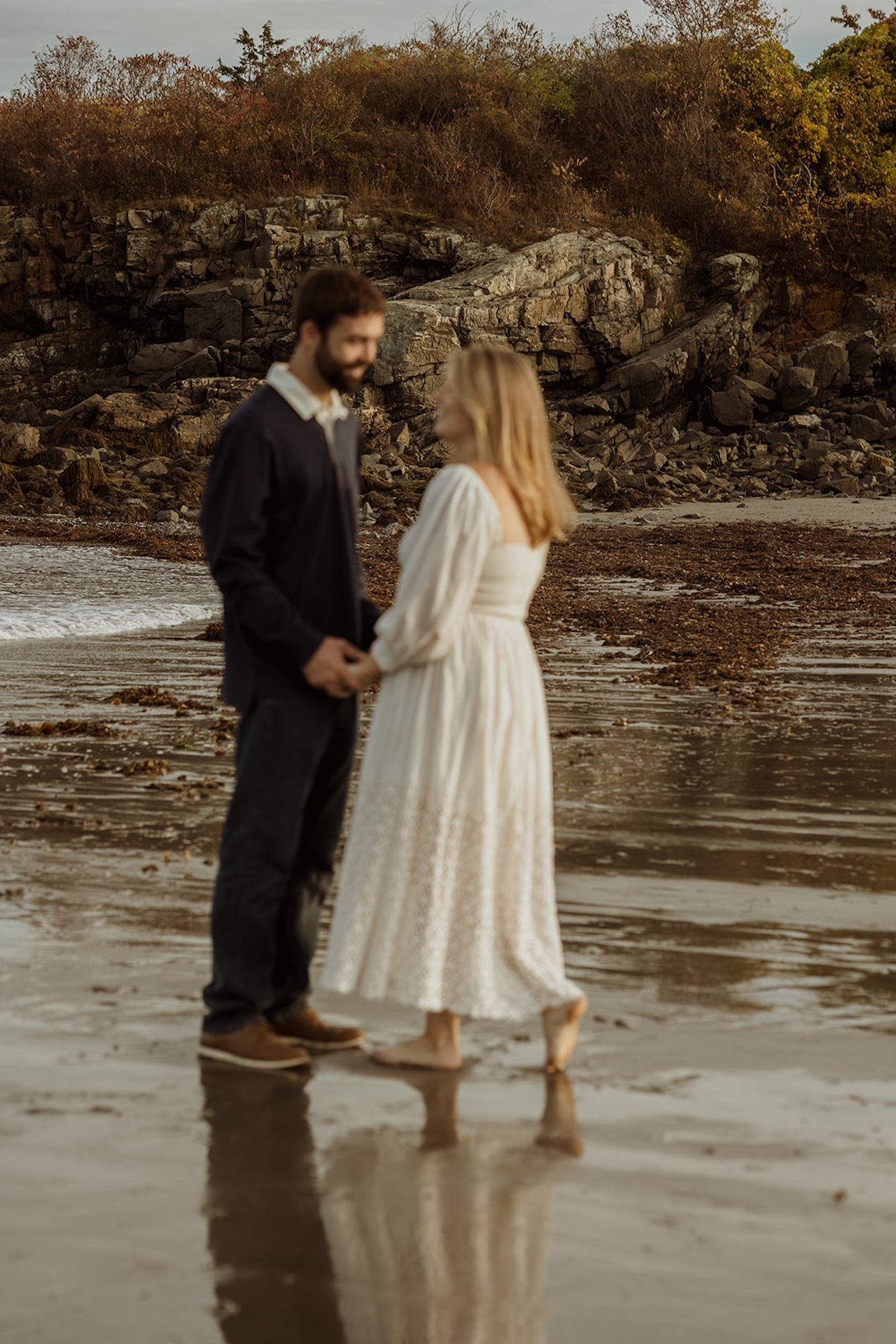 Romantic sunset engagement photo of a couple dancing barefoot in the sand near Nubble Lighthouse in York Maine with ocean waves behind them.