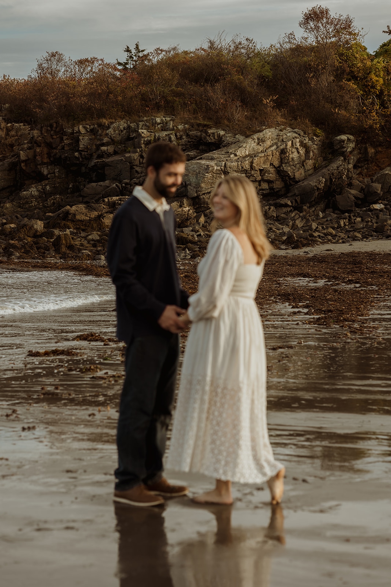 Romantic engagement photo of couple dancing barefoot in the sand near Nubble Lighthouse in York Maine with ocean waves behind them.