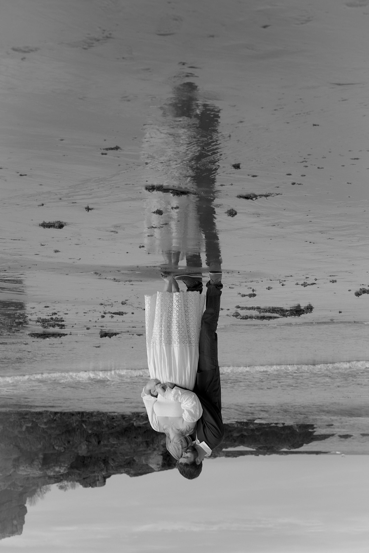 Artistic reflection photo of couple embracing in wet sand at Nubble Lighthouse in York Maine with mirrored ocean water beneath them.