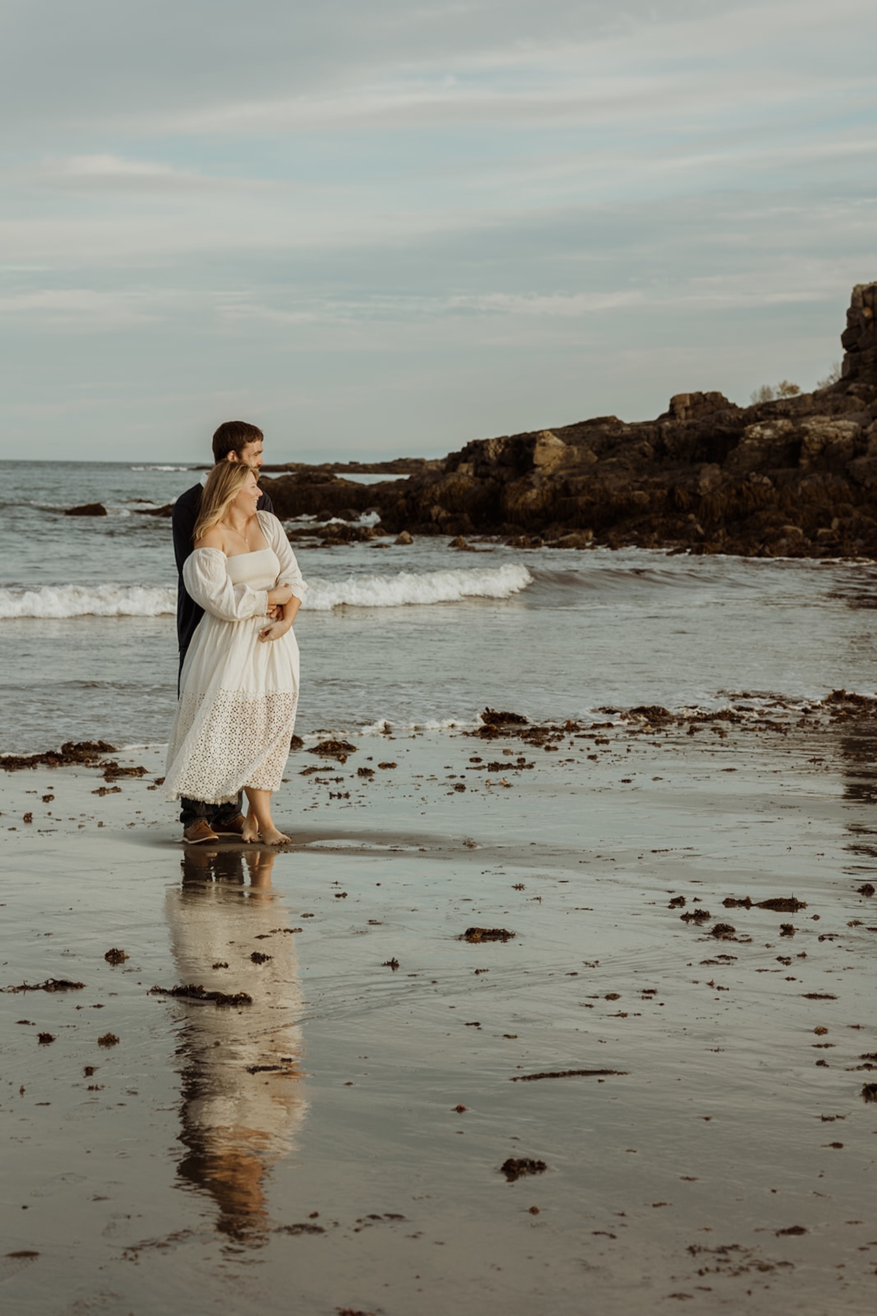 Scenic coastal engagement portrait of couple standing together on the beach near Nubble Lighthouse in York Maine with rocky cliffs behind them.