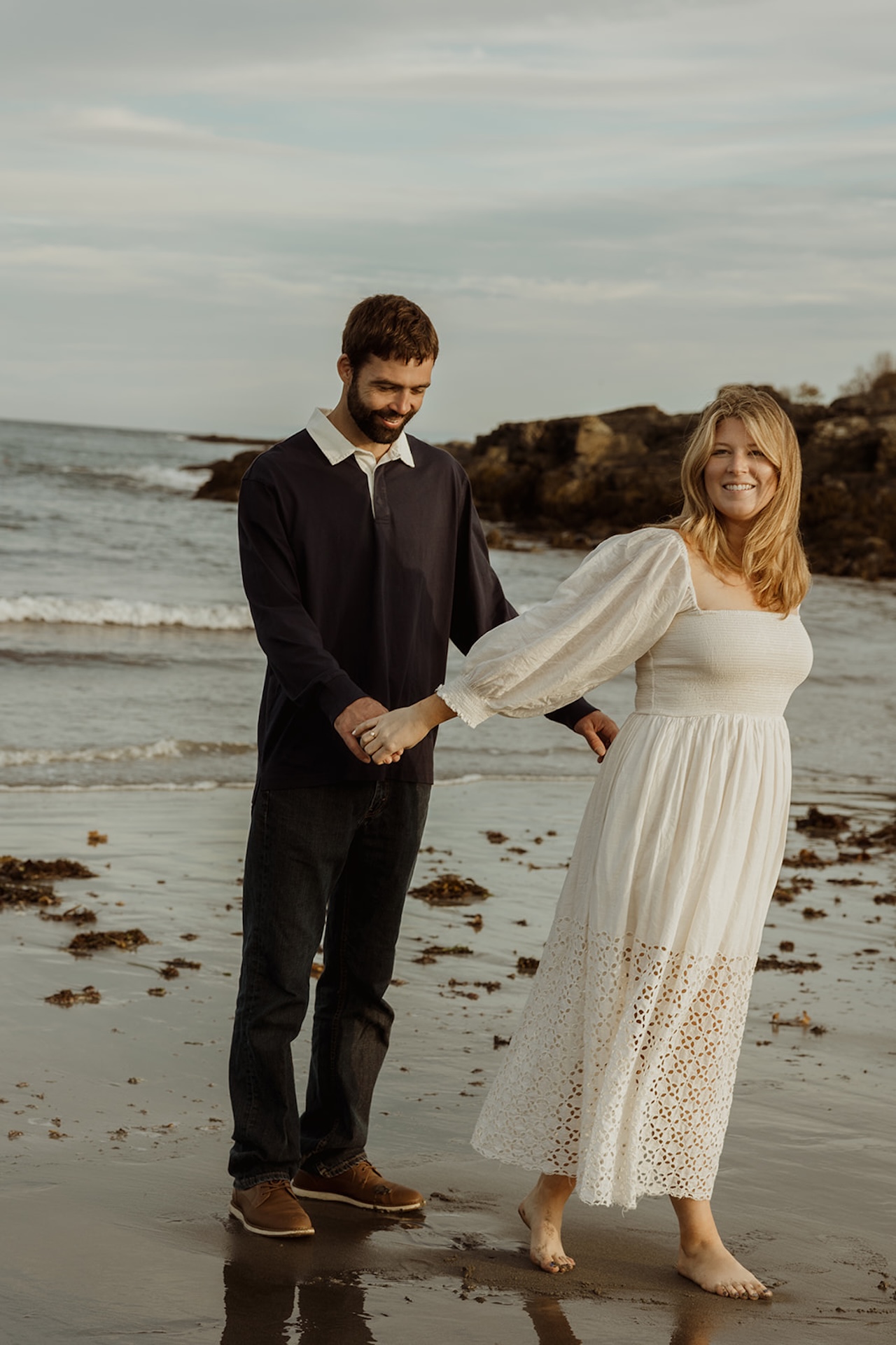 Wide engagement photo of couple dancing barefoot in the sand near Nubble Lighthouse in York Maine with ocean waves behind them.