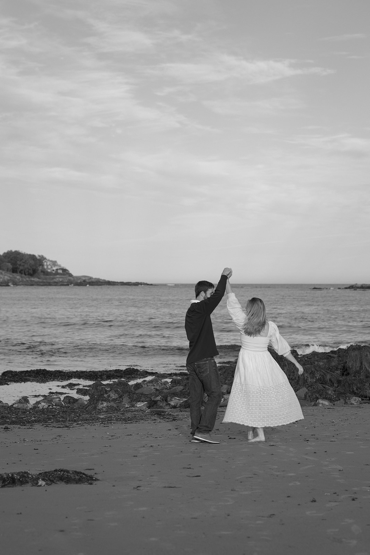 Black and white engagement photo of couple twirling on the beach with rocky shoreline and open sky in the background.
