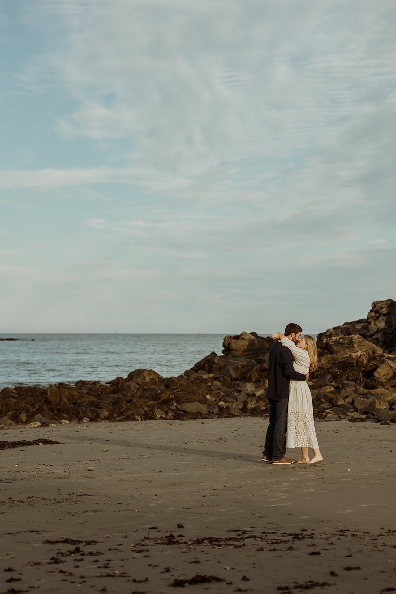 Romantic engagement photo of a couple hugging and sharing a kiss on the sand near Nubble Lighthouse in York Maine with ocean waves behind them.