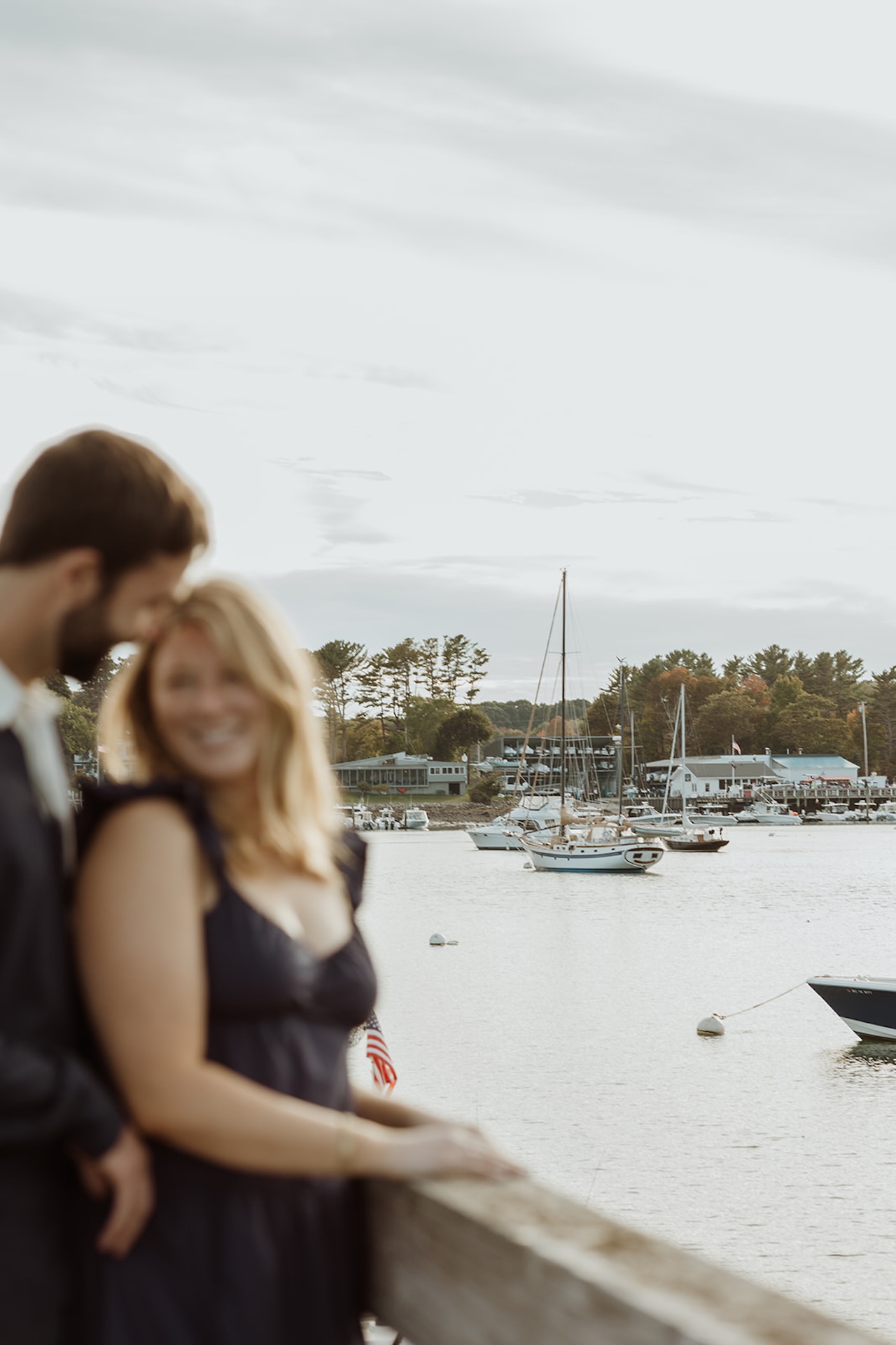 Soft, romantic engagement photo at Stage Neck Inn (their wedding venue) with sailboats in the harbor and the couple leaning together along the railing.