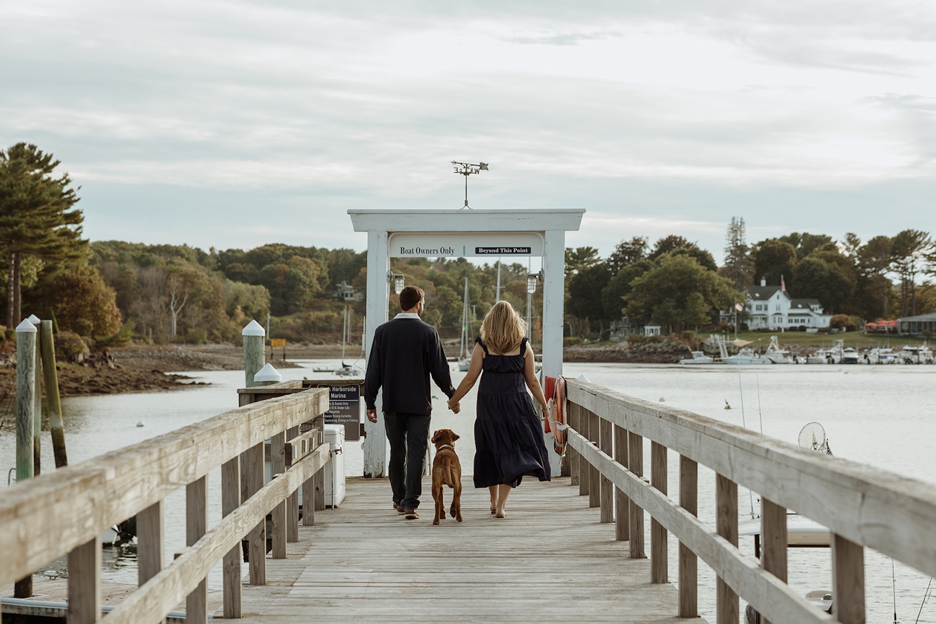 Couple walking hand in hand with their dog down a wooden dock at a quiet coastal marina during golden hour engagement photos.