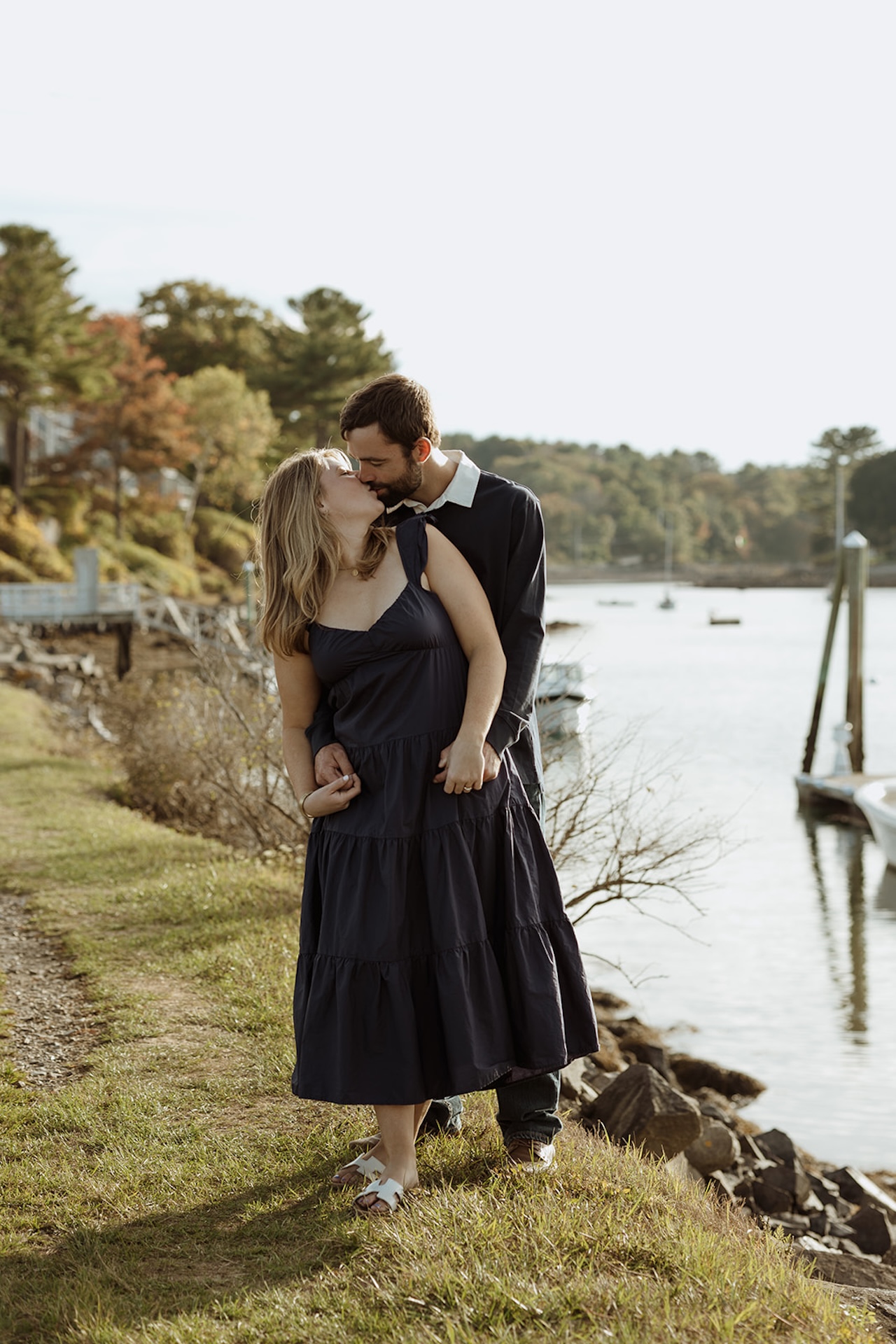Intimate engagement portrait of couple kissing along the shoreline near Nubble Lighthouse in York Maine with calm harbor water behind them.