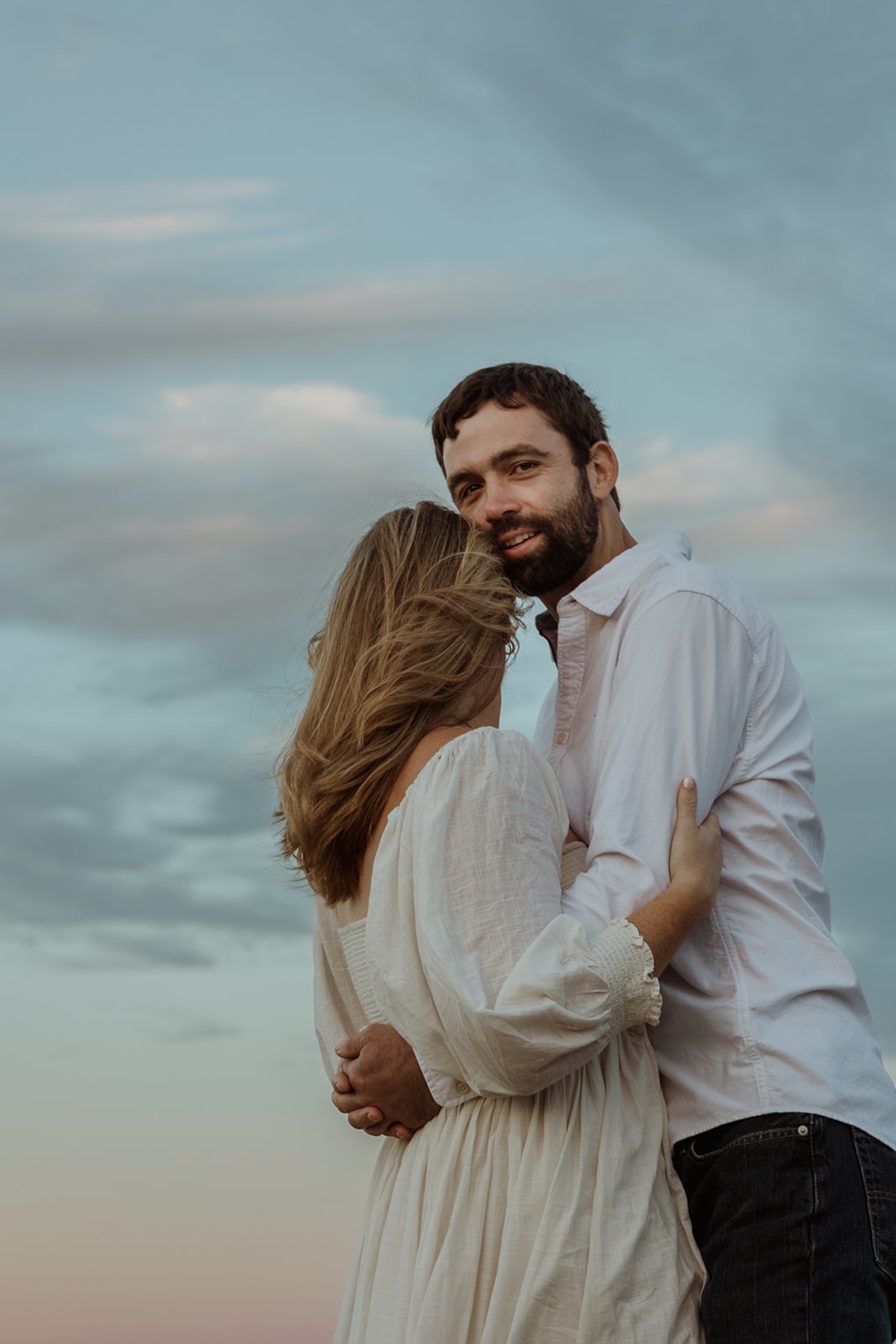 Close engagement portrait of couple embracing with wind blowing through her hair under a pastel sunset sky.