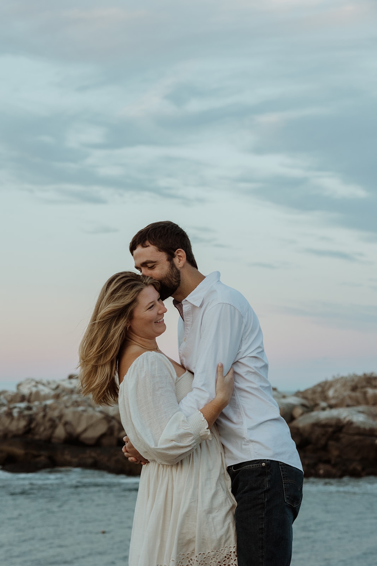 Romantic candid engagement portrait of couple laughing together while embracing on rocky beach coastline.
