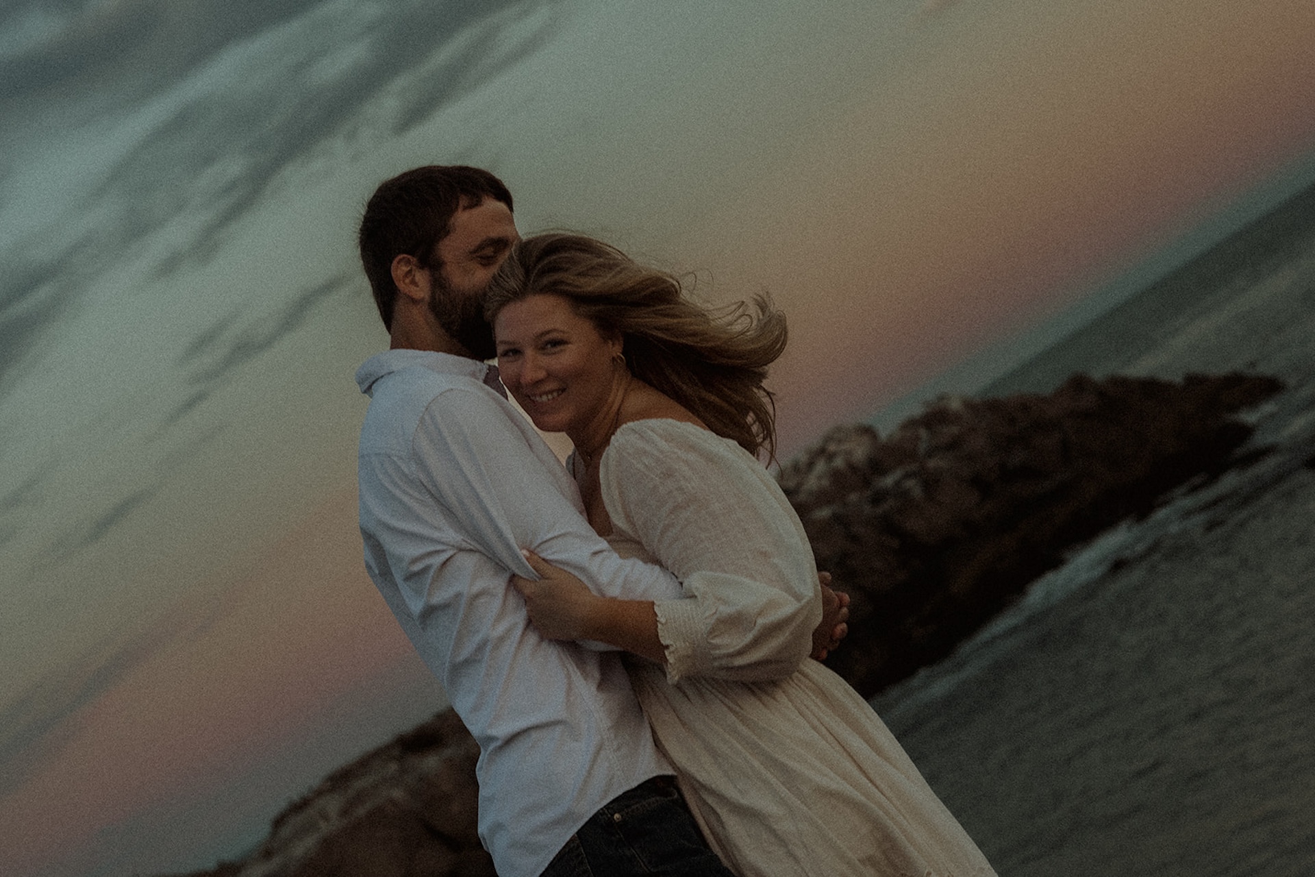 Wind-swept engagement portrait of couple hugging on rocky coastline at sunset with dramatic ocean backdrop.