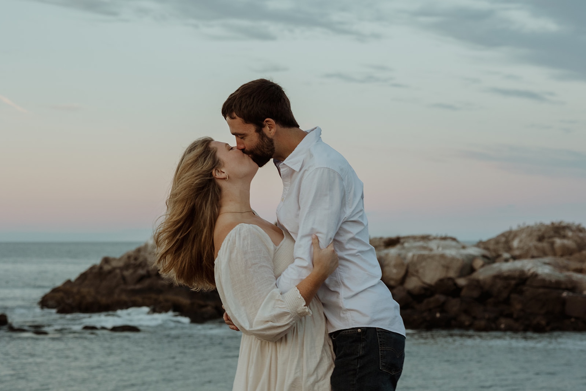 Wind-swept engagement portrait of couple kissing on rocky coastline at sunset with dramatic ocean backdrop near Nubble Lighthouse in York Maine