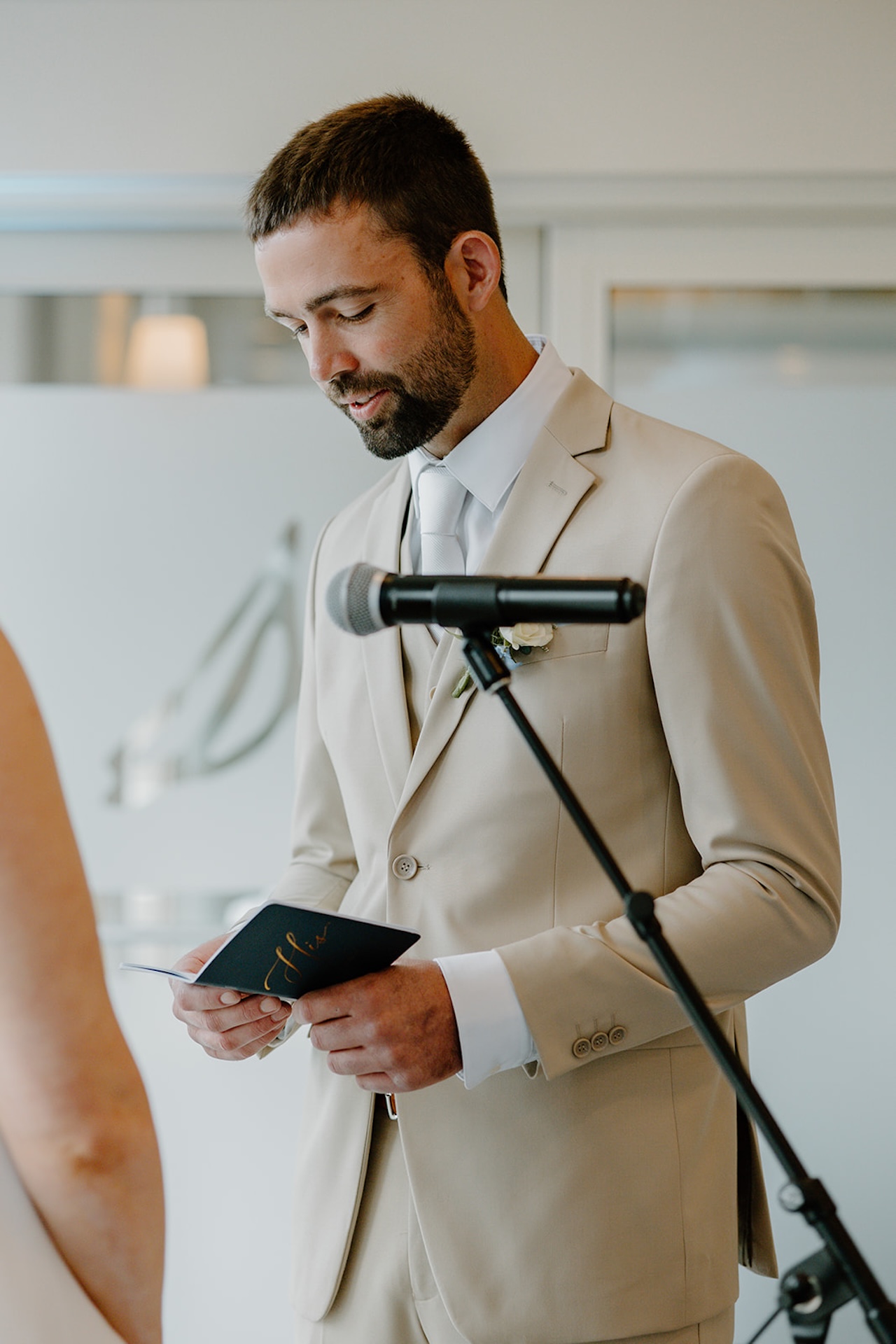 Groom reading handwritten vows at the ceremony microphone wearing a tan wedding suit.